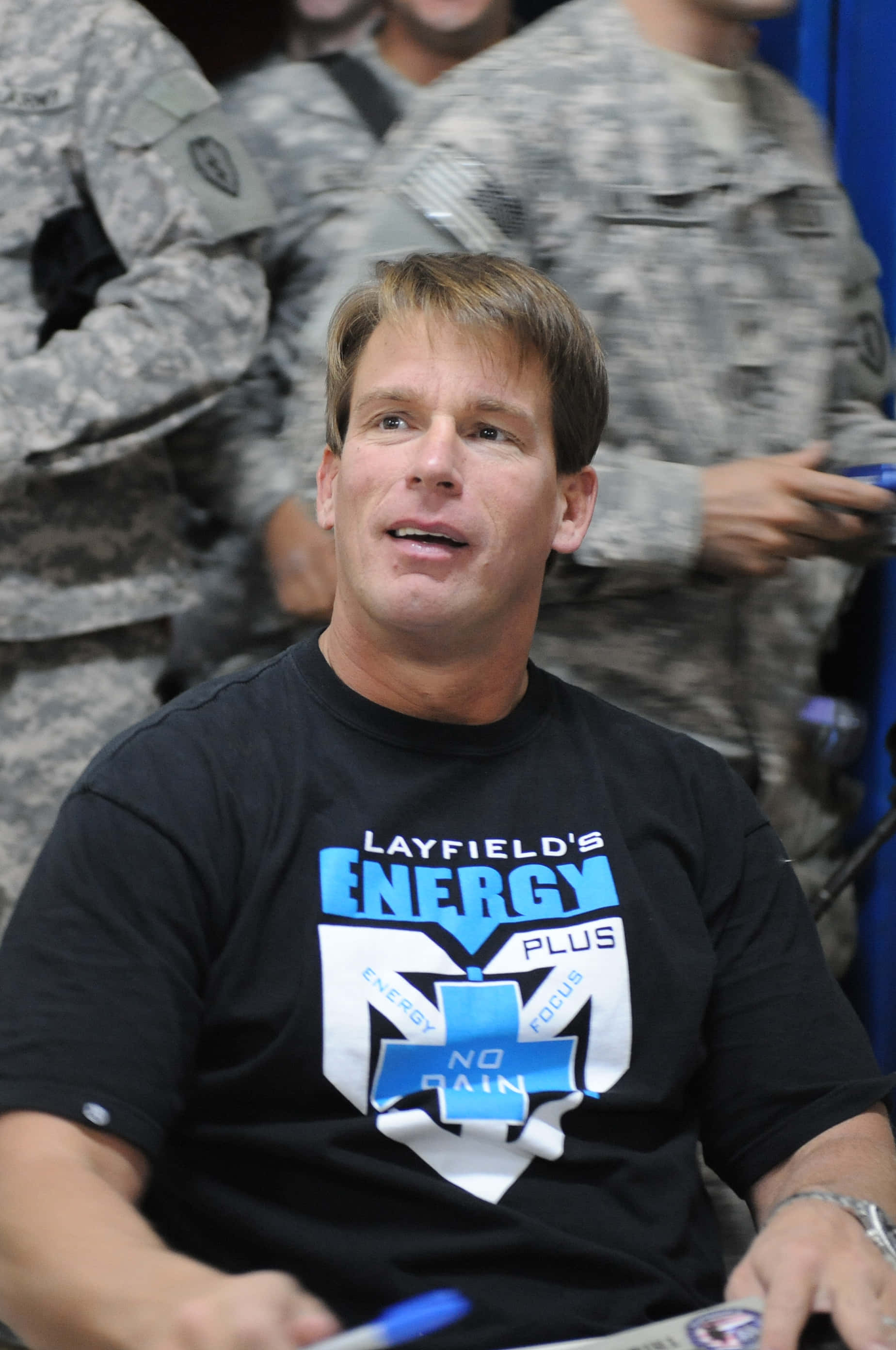 WWE 桌布 壁紙 — 一名身穿印有「Layfield's Energy Plus」圖案黑色T恤的男子，坐在模糊的迷彩軍人前面。