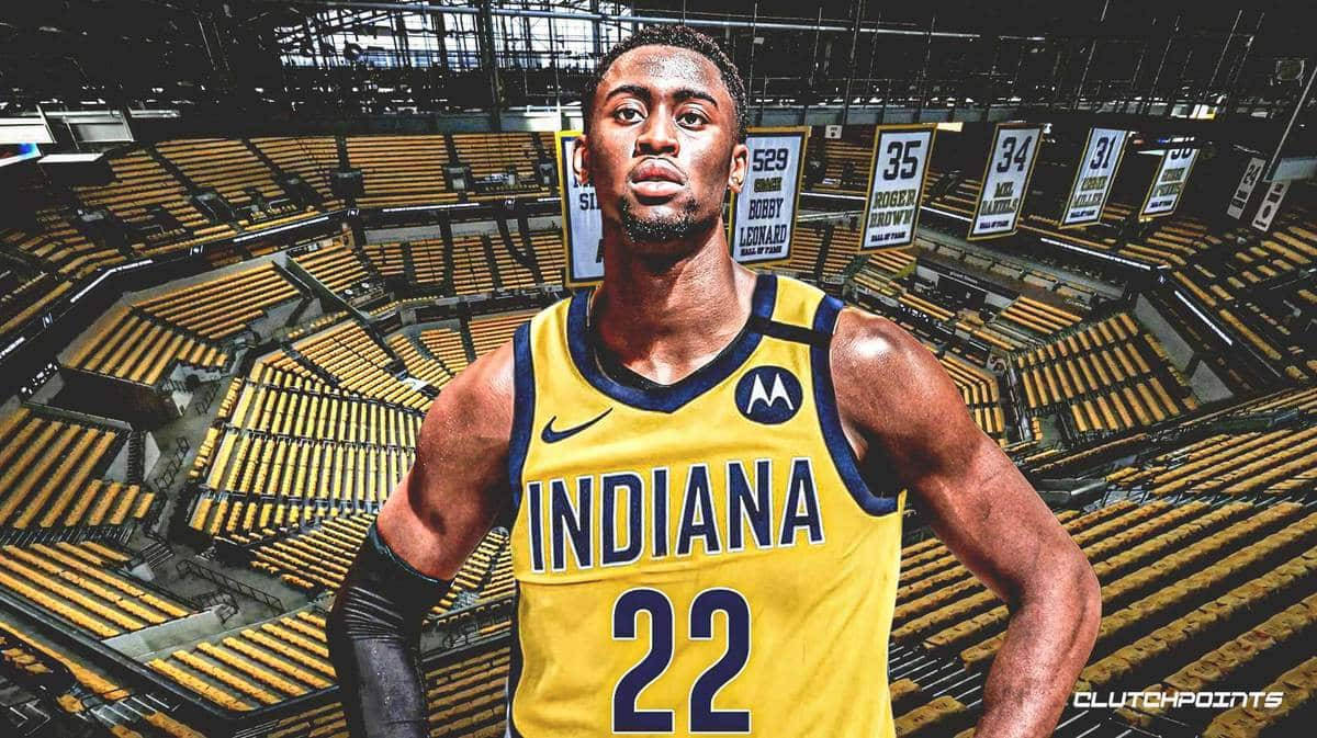 caris levert 桌布 壁紙 — 一名身穿印第安納球衣的籃球運動員自信地站在一個空蕩蕩的體育館內，周圍有黃色座位和退役號碼橫幅。
