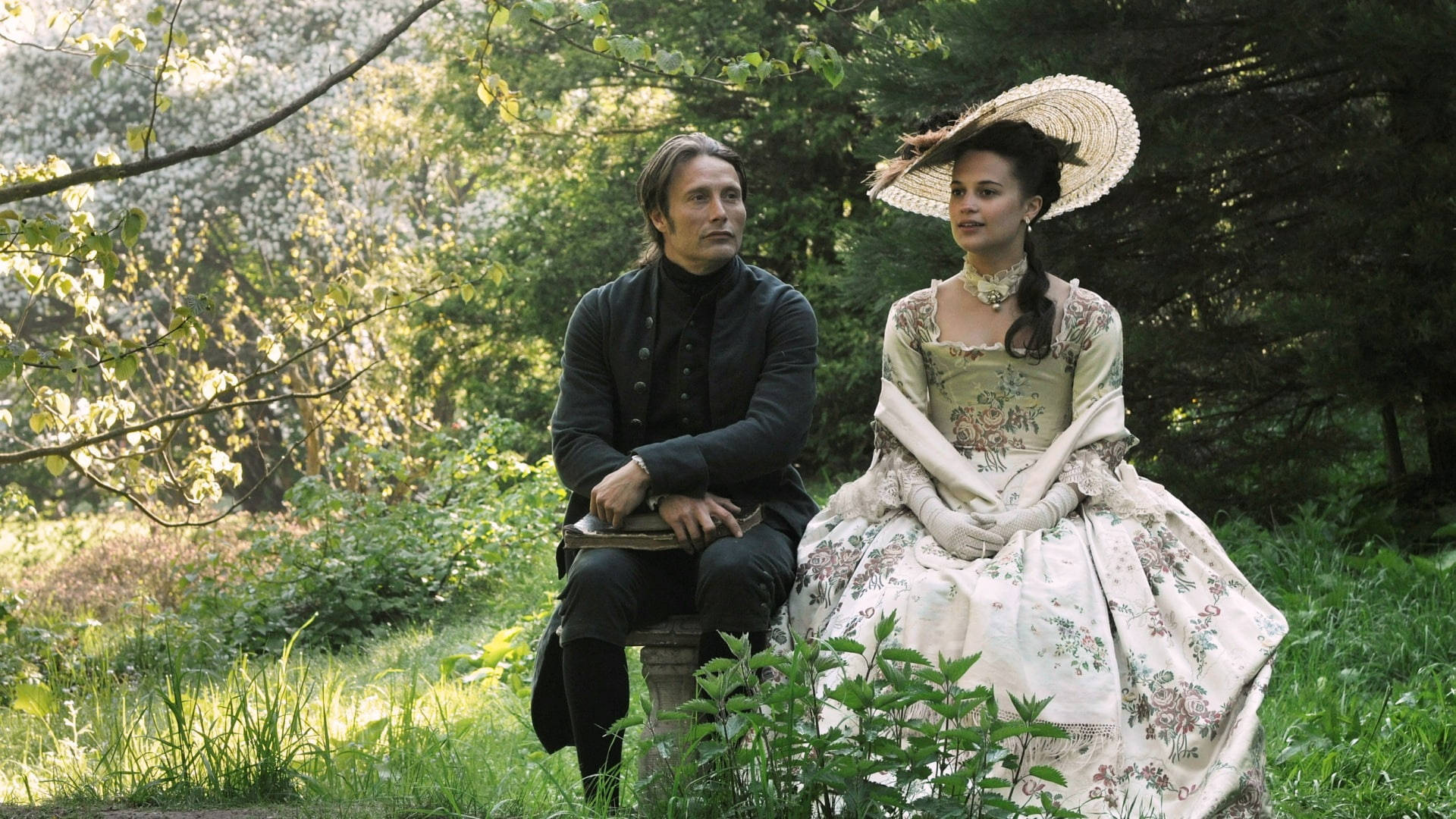 Mads Mikkelsen 桌布 壁紙 — 一名身穿古裝的男子和女子坐在陽光普照的花園中，周圍環繞著綠樹和盛開的樹木。