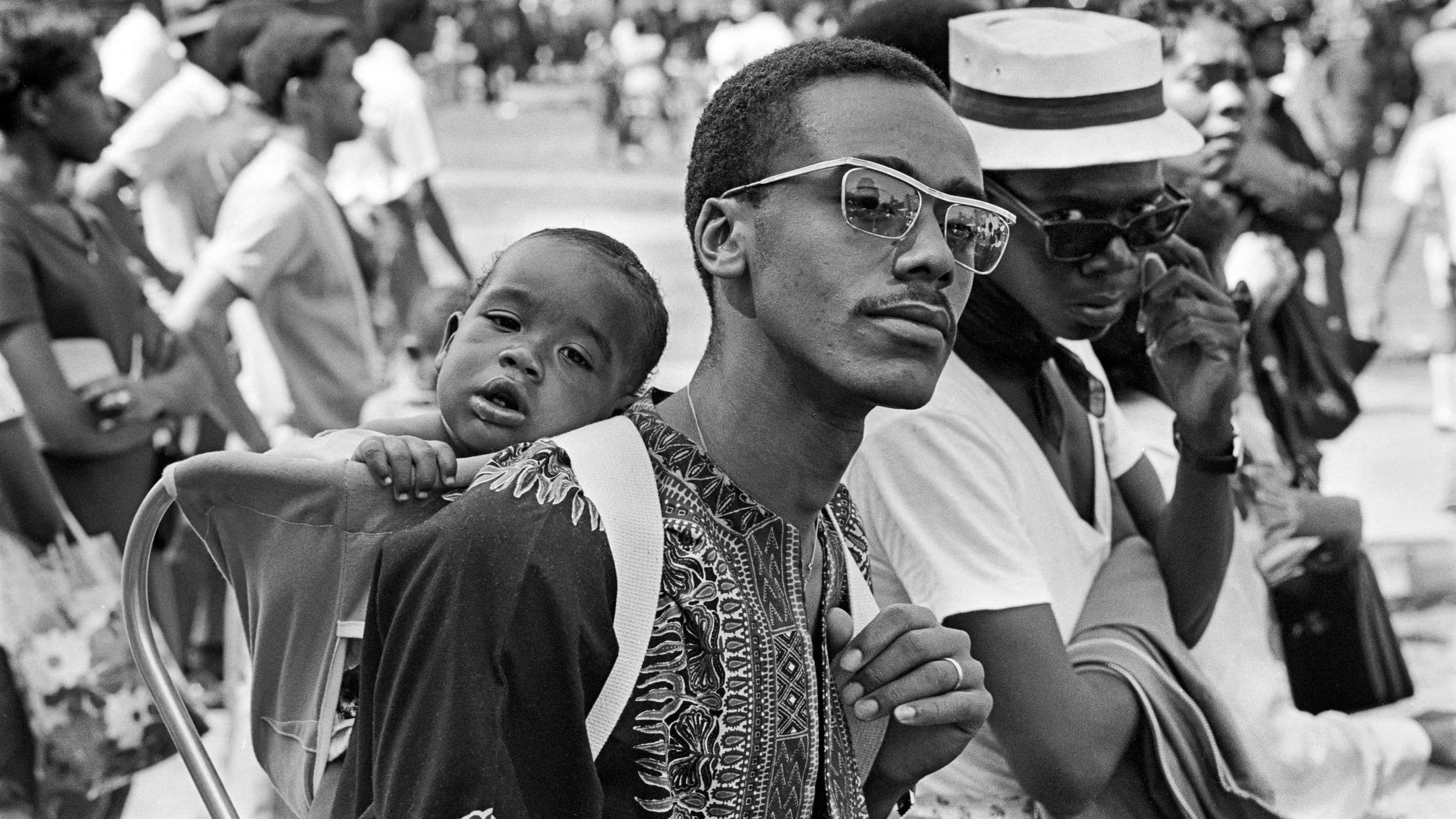 Black Culture 桌布 壁紙 — 一名身穿圖案襯衫、戴眼鏡的男子，在戶外坐著，肩上扛著一個孩子，身後是模糊的人群。