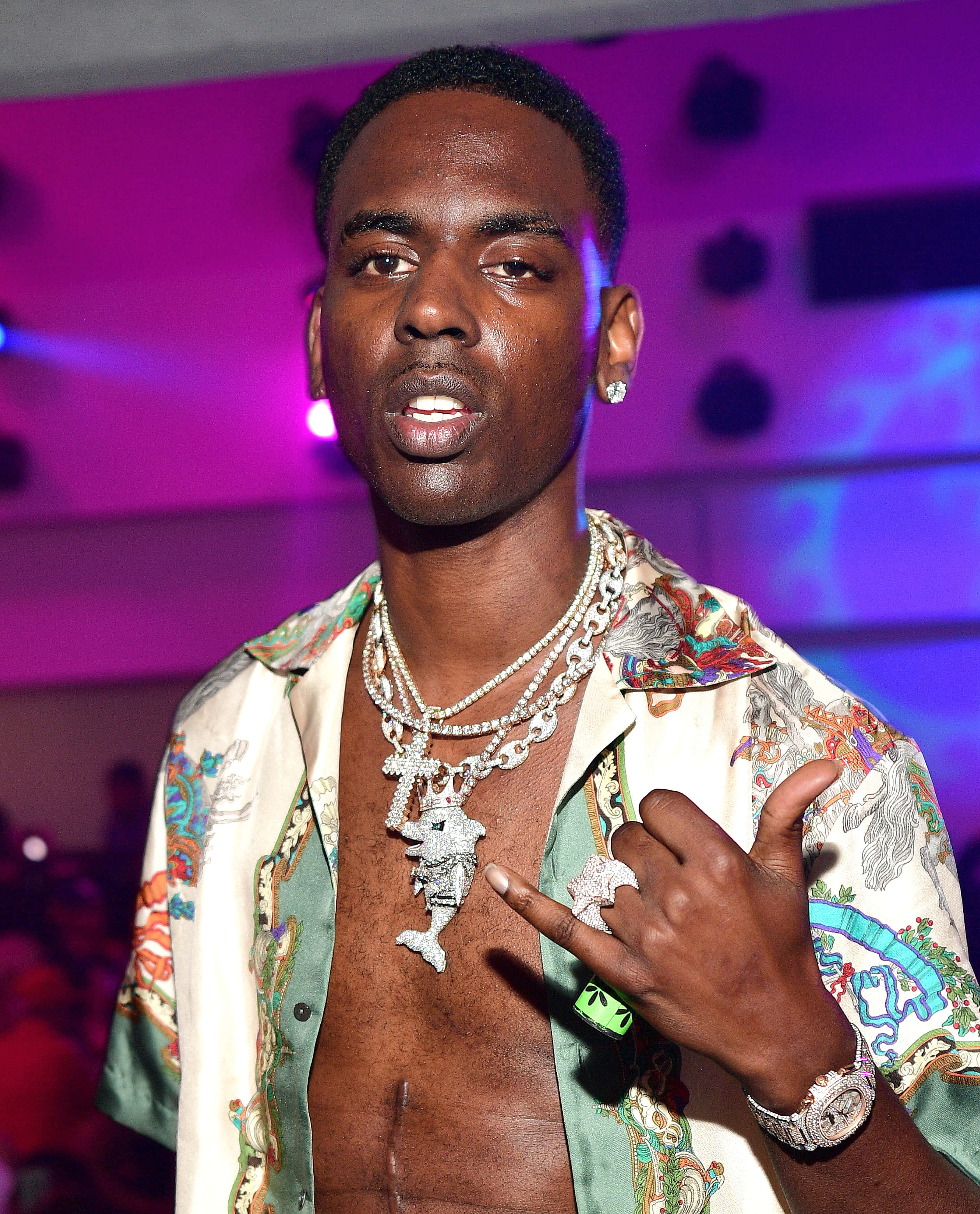 Young Dolph 桌布 壁紙 — 一名身穿圖案襯衫和鑽石珠寶的男子，在充滿活力的紫色燈光派對上自信地擺姿勢。