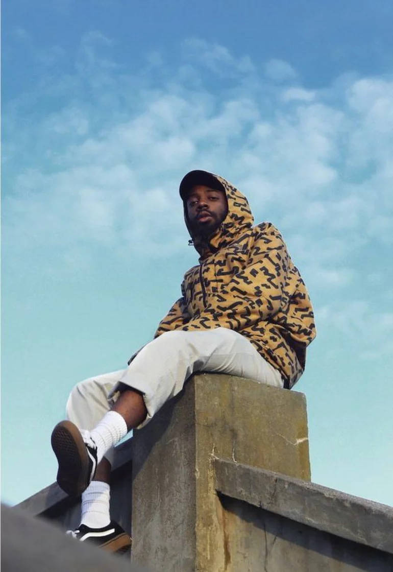 Brent Faiyaz 桌布 壁紙 — 一名身穿圖案連帽上衣的年輕男子坐在混凝土結構上，背景是晴朗的藍天和白雲。
