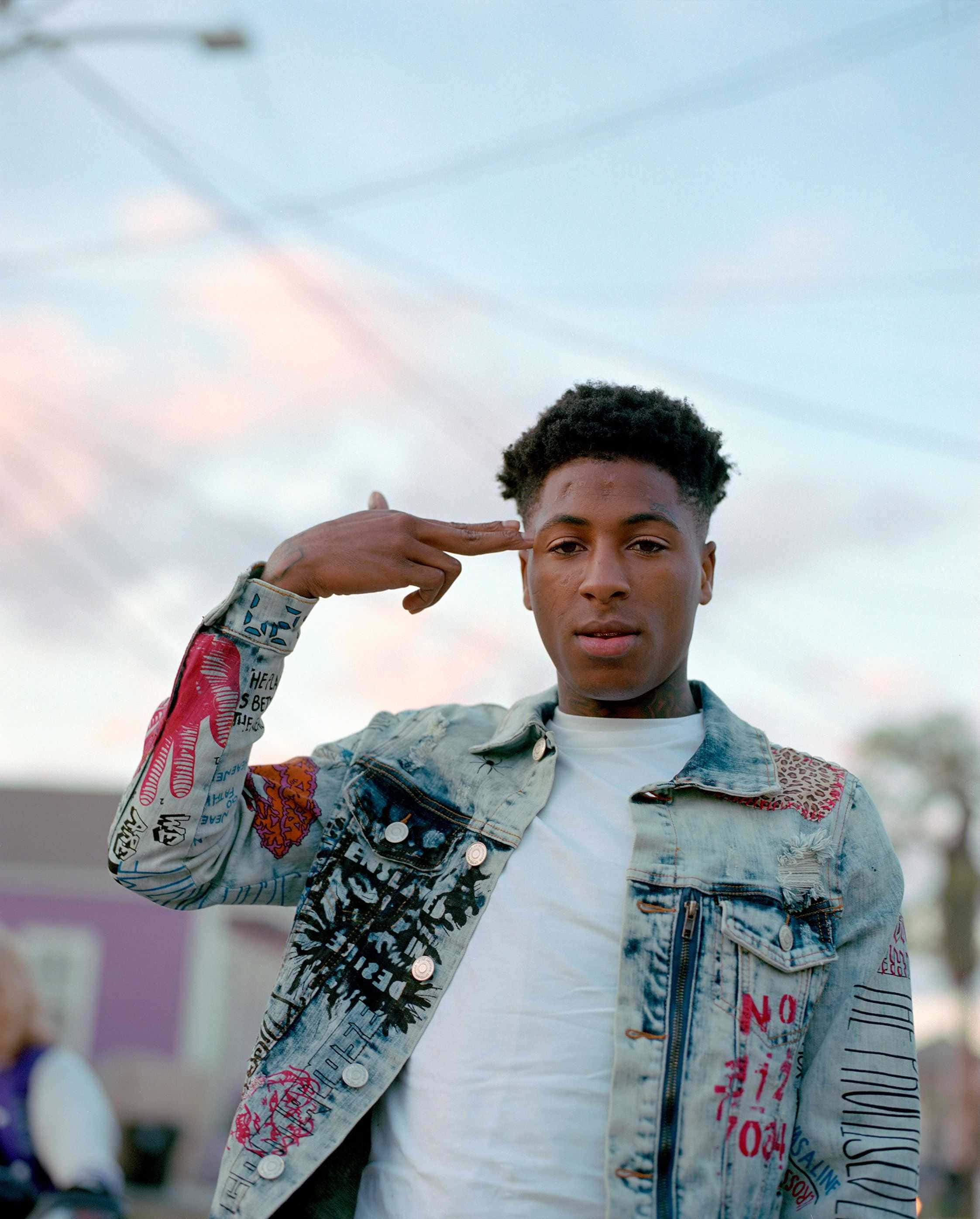 NBA YoungBoy 桌布 壁紙 — 一名身穿塗鴉牛仔外套的年輕男子，在柔和的暮色天空下以嚴肅的眼神敬禮。