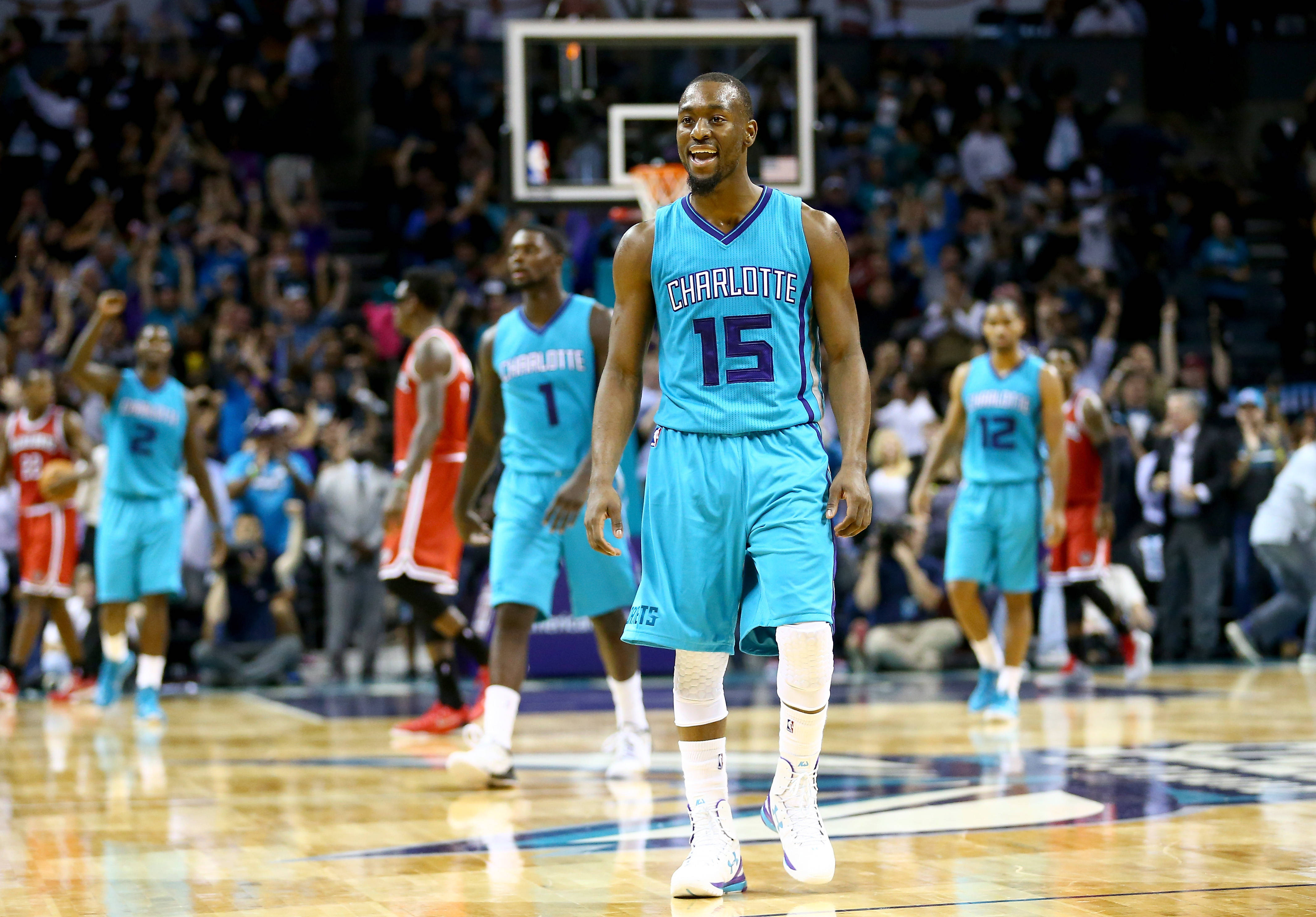 Kemba Walker 桌布 壁紙 — 一名身穿夏洛特球衣的籃球運動員在光亮的球場上自信微笑，身後有隊友和歡呼的觀眾。