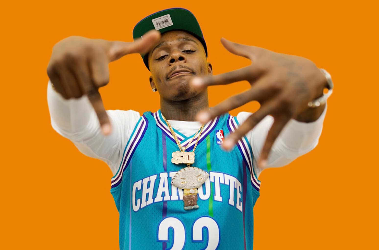 Dababy 桌布 壁紙 — 一名身穿夏洛特籃球球衣的男子在純橙色背景前自信地擺姿勢，雙手指向自己的臉。
