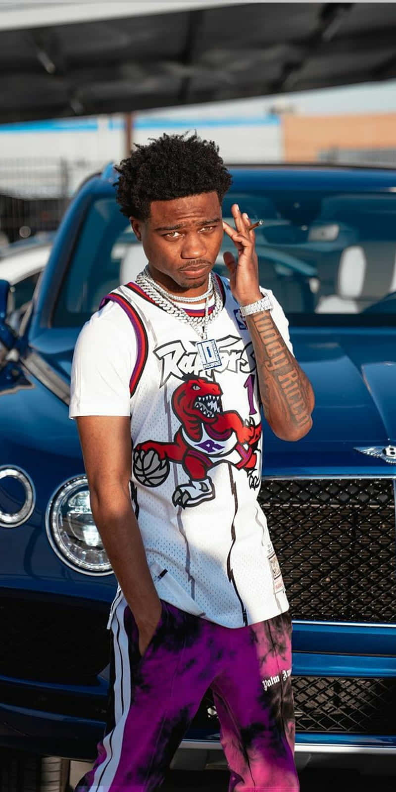 Roddy Ricch 桌布 壁紙 — 一名身穿多倫多暴龍隊球衣和紫色紮染褲的年輕男子，自信地在一輛藍色豪華車旁擺姿勢。