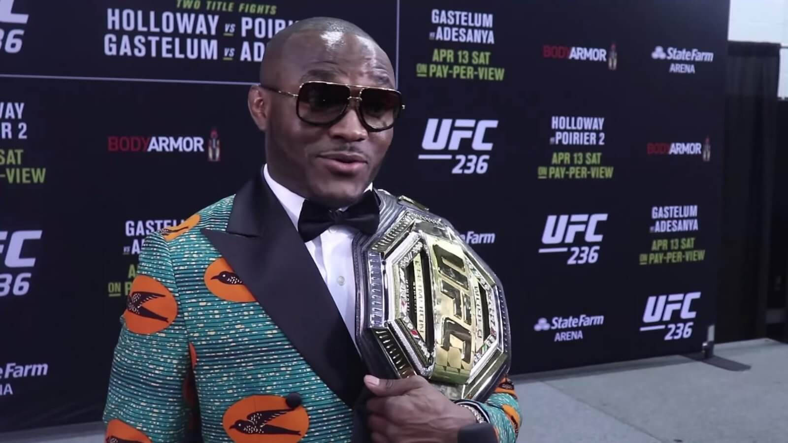 UFC 桌布 壁紙 — 一名身穿大膽圖案西裝和太陽眼鏡的拳手在記者會上手持UFC冠軍腰帶。
