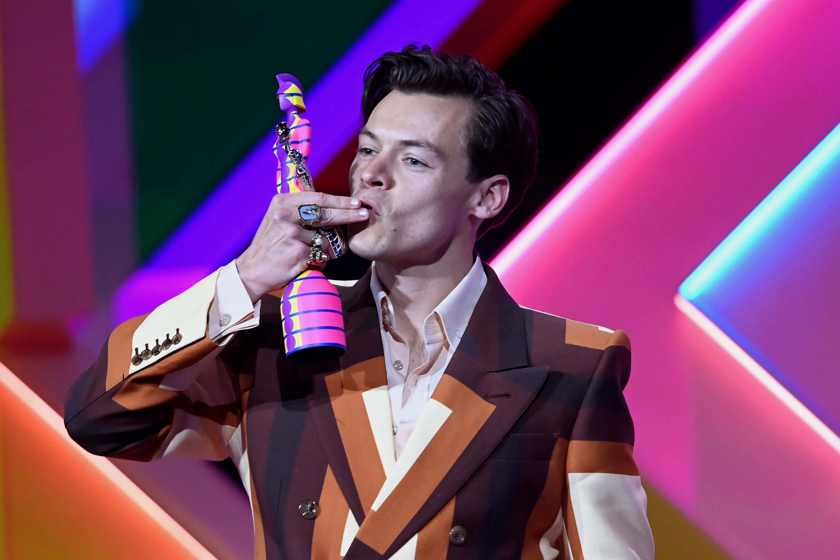Harry Styles 2021 桌布 壁紙 — 一名身穿大膽幾何圖案西裝的男子，在充滿活力的霓虹燈舞台背景下親吻一個色彩繽紛的獎盃。