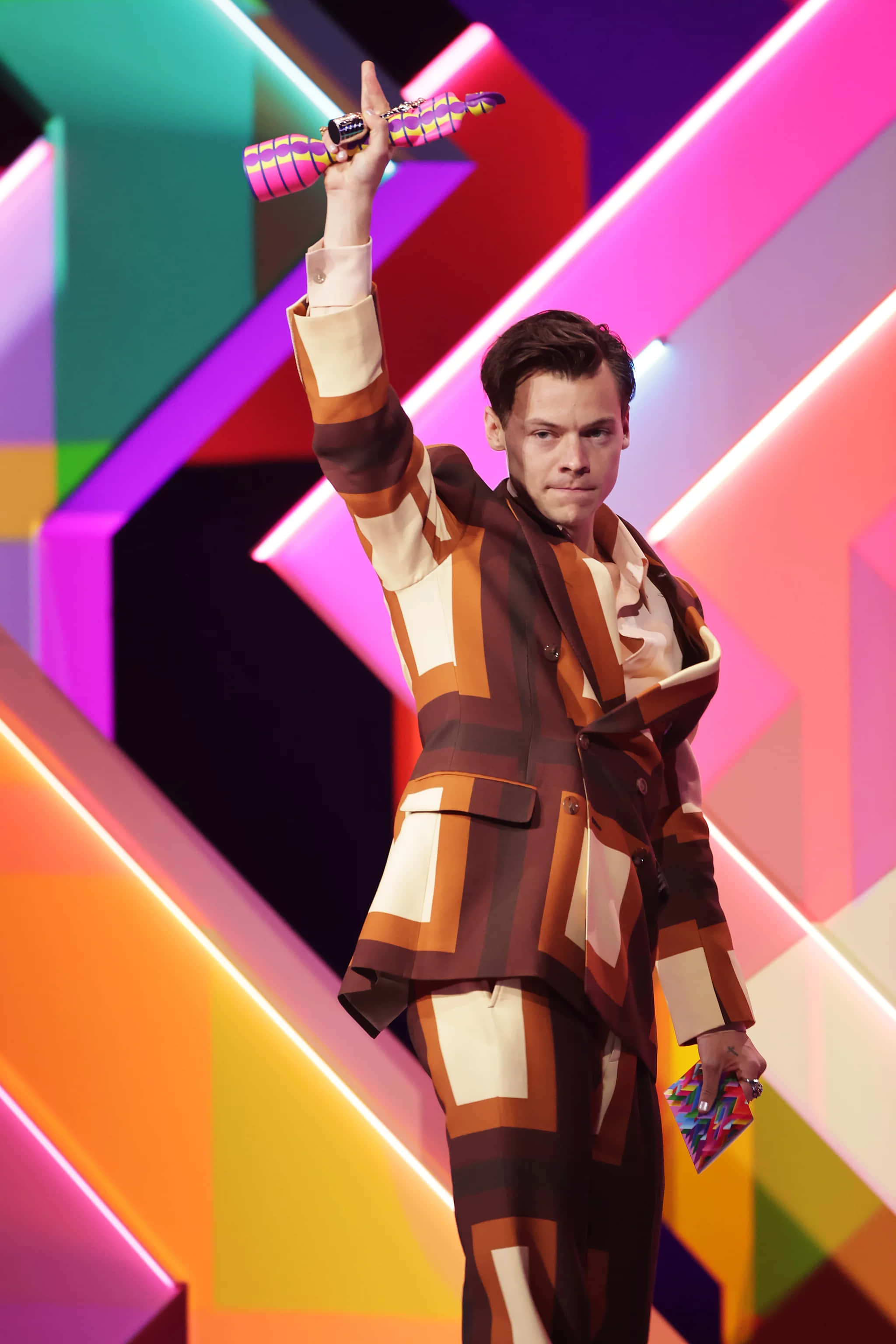 Harry Styles 2021 桌布 壁紙 — 一名身穿大膽幾何圖案西裝的男子，在充滿活力的霓虹燈舞台背景下高舉色彩繽紛的獎項。