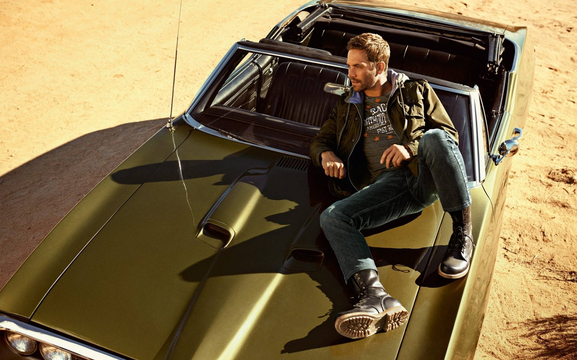 Paul Walker 桌布 壁紙 — 一名身穿夾克和靴子的男子，悠閒地躺在一輛停在陽光普照的沙漠中的復古橄欖綠色肌肉車上。