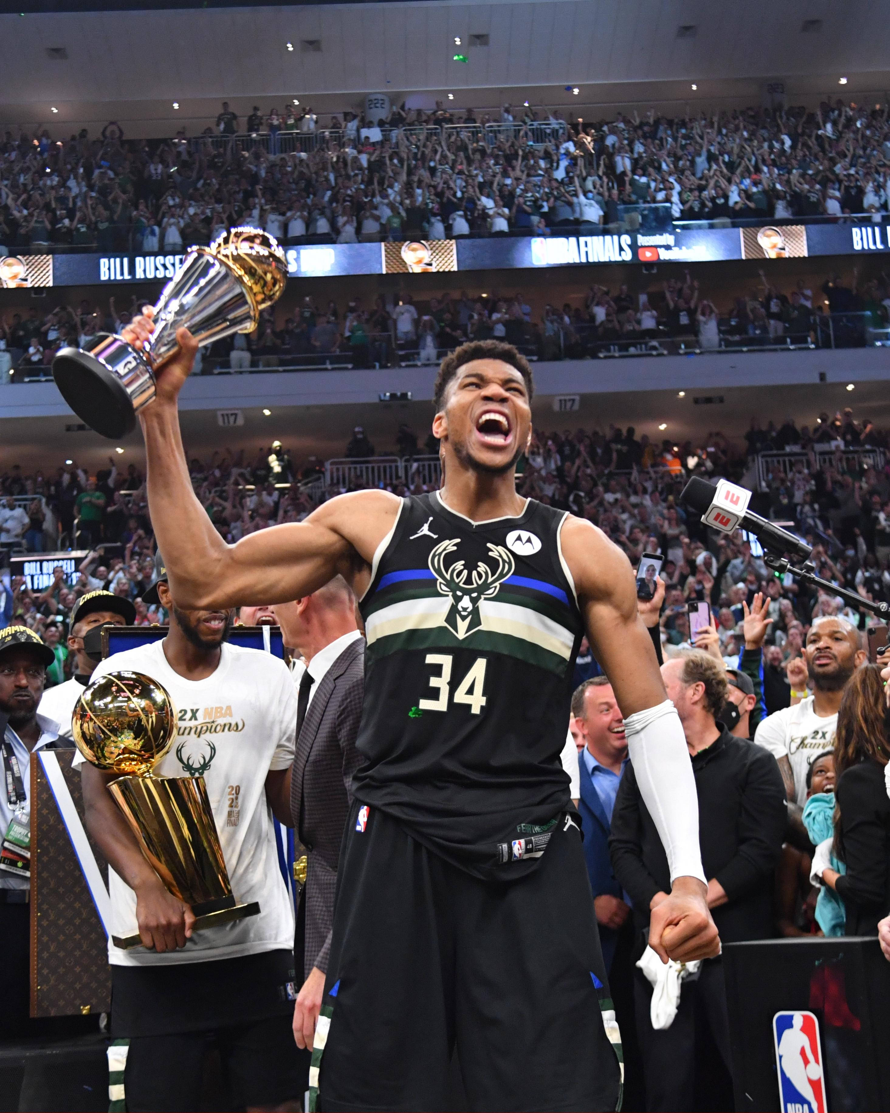 Giannis 桌布 壁紙 — 一名身穿密爾瓦基公鹿隊球衣的籃球運動員在歡呼的人群面前咆哮著，勝利地高舉NBA總冠軍獎盃。