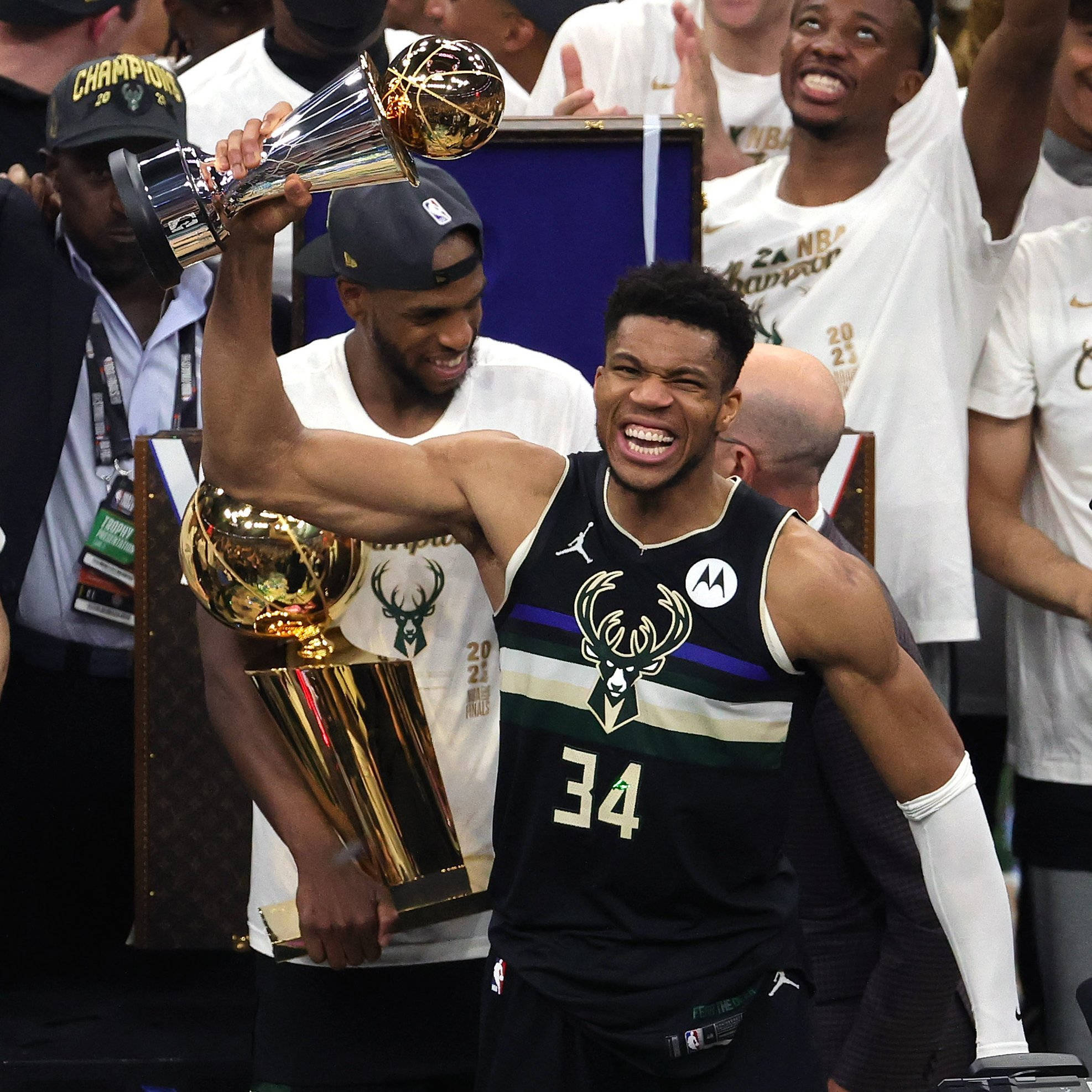 Giannis 桌布 壁紙 — 一名身穿密爾瓦基公鹿隊球衣的籃球運動員高舉NBA總冠軍獎盃，隊友們在他周圍慶祝。