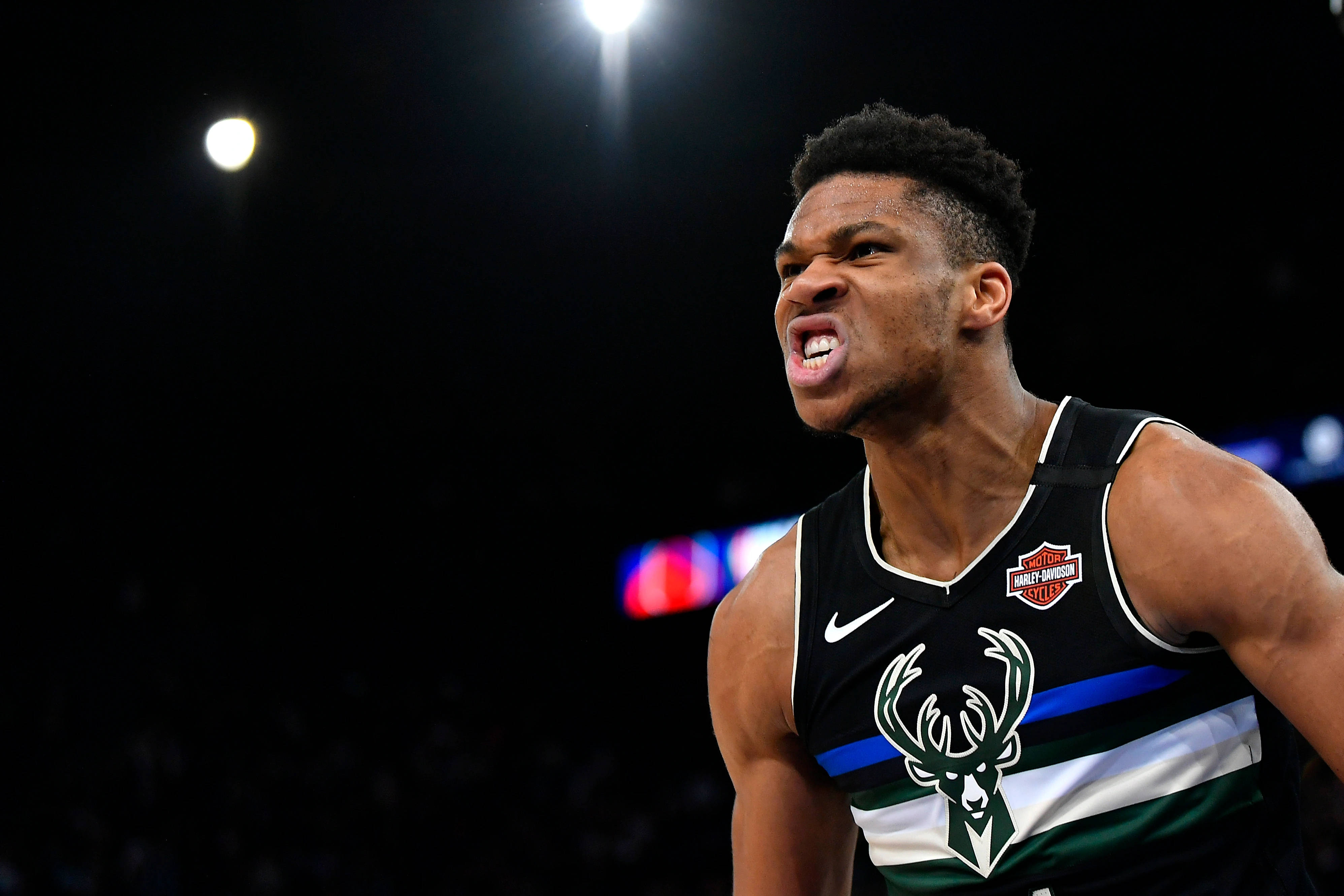 Giannis 桌布 壁紙 — 一名身穿密爾瓦基公鹿隊球衣的籃球運動員在黑暗背景的場館燈光下激烈咆哮。