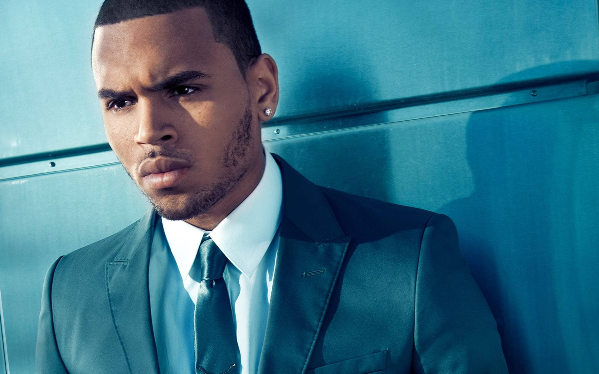 Chris Brown 桌布 壁紙 — 一名身穿尖挺青綠色西裝的男子，在戲劇性的燈光下，以銳利的目光靠著藍色金屬牆。