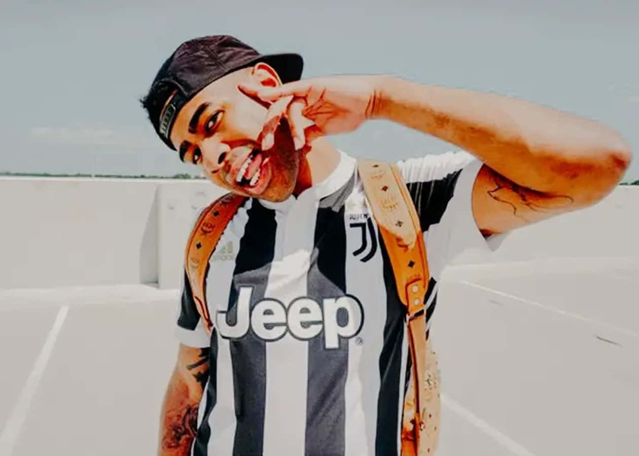 Prettyboyfredo 桌布 壁紙 — 一名身穿尤文圖斯球衣的男子在晴朗天空下的明亮屋頂上微笑並做出和平手勢。