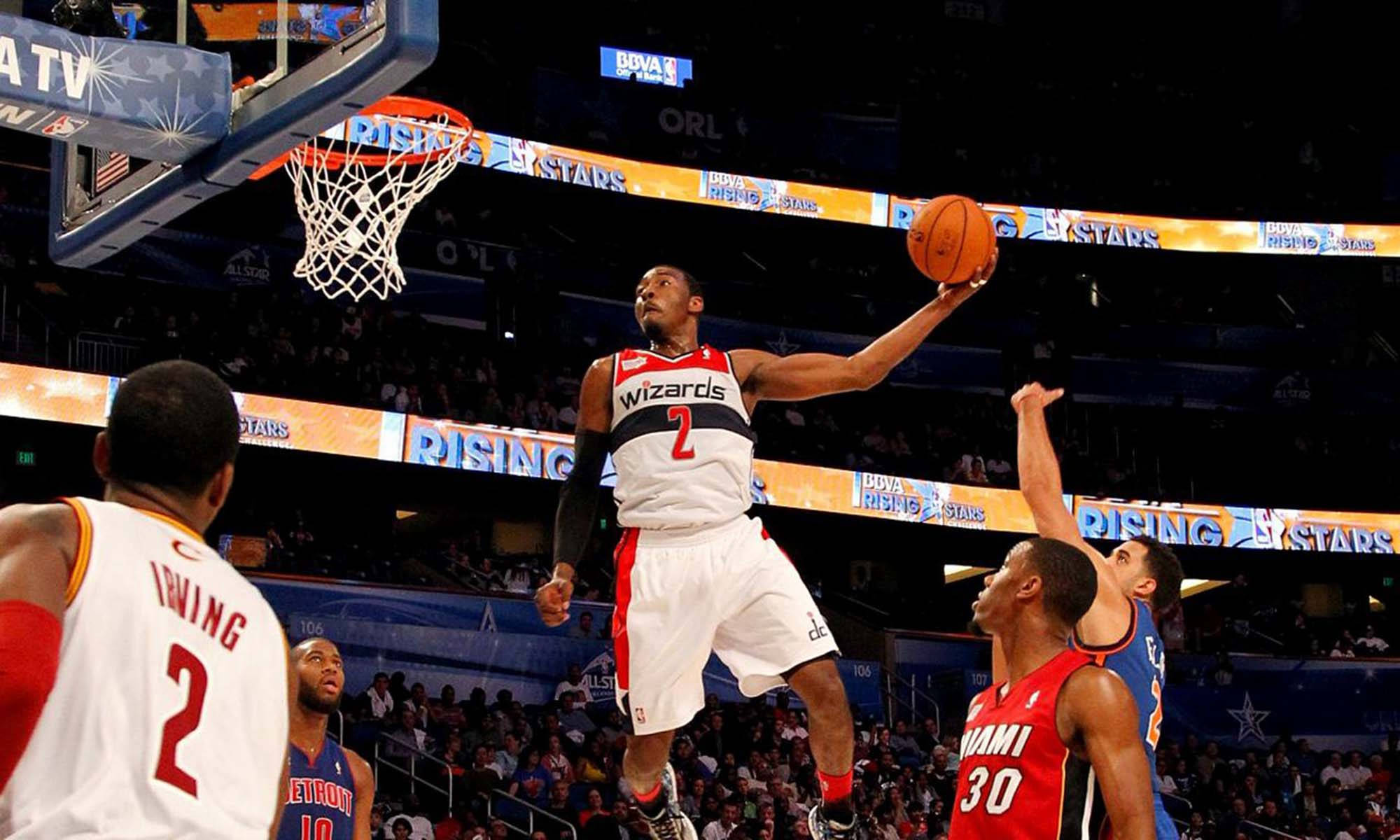 John Wall 桌布 壁紙 — 一名身穿巫師隊球衣的籃球運動員在場館燈光下高高躍起灌籃，防守球員在下方觀望。