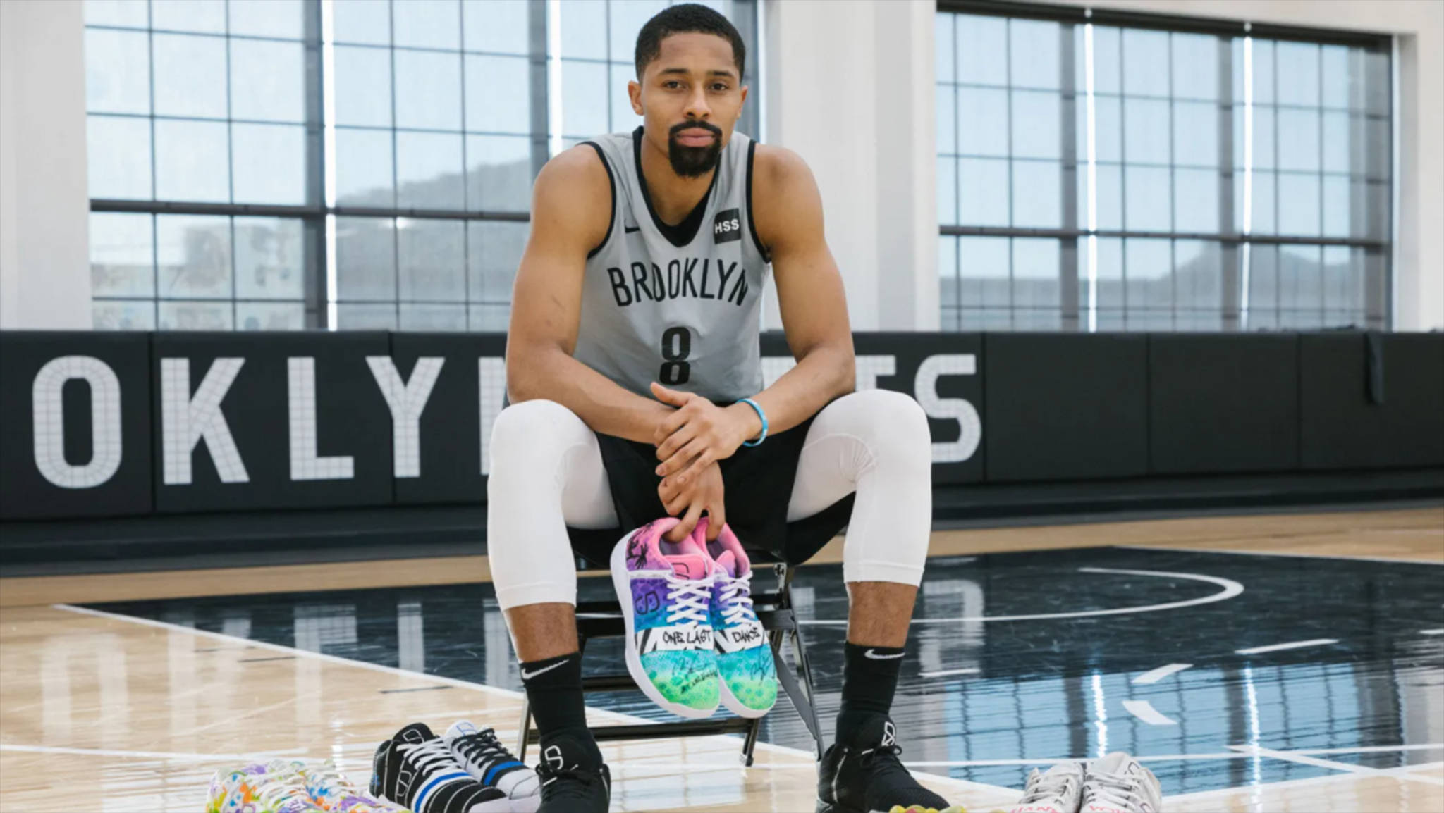 spencer dinwiddie 桌布 壁紙 — 一名身穿布魯克林球衣的籃球運動員坐在球場上，手持色彩繽紛的運動鞋，附近還有其他鞋子。