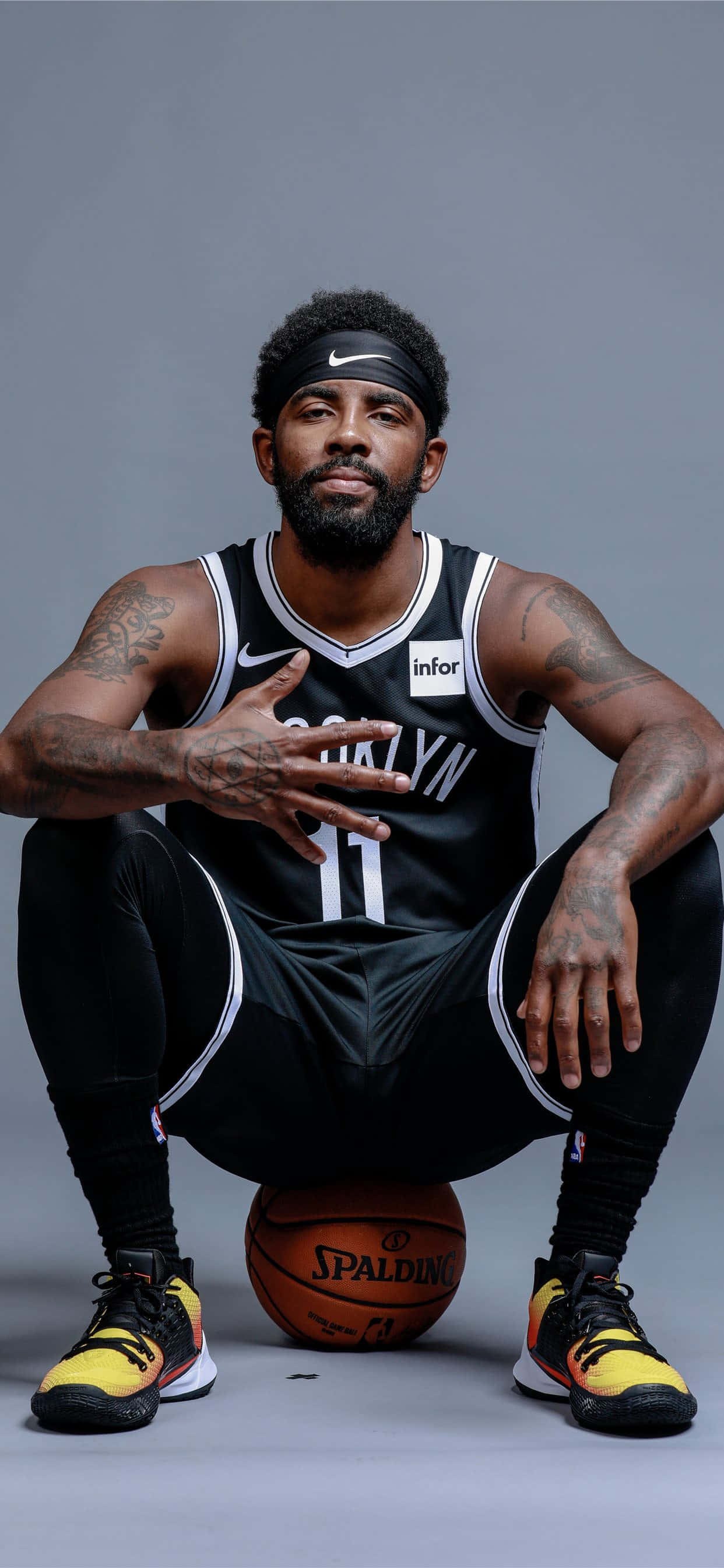 Kyrie Irving Nets 桌布 壁紙 — 一名身穿布魯克林球衣的籃球運動員自信地蹲下，雙腿之間夾著一個Spalding籃球，背景是純灰色的攝影棚。