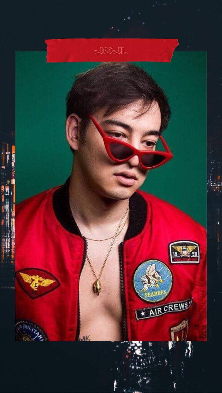 joji 桌布 壁紙 — 一名身穿帶有補丁的紅色飛行員夾克和紅色太陽眼鏡的男子，在帶有城市景觀倒影的綠色背景前擺姿勢。