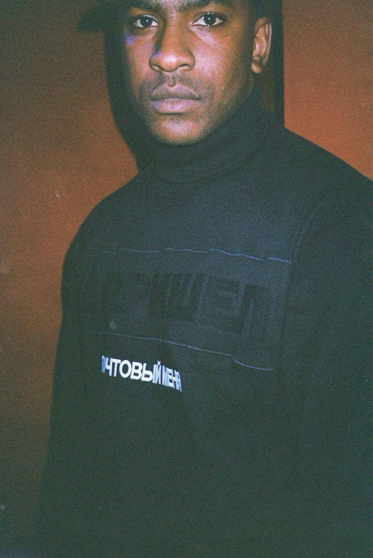 Skepta 桌布 壁紙 — 一名身穿帶有西里爾文字的黑色高領毛衣的年輕男子，在暖色調的牆壁前凝視著鏡頭。