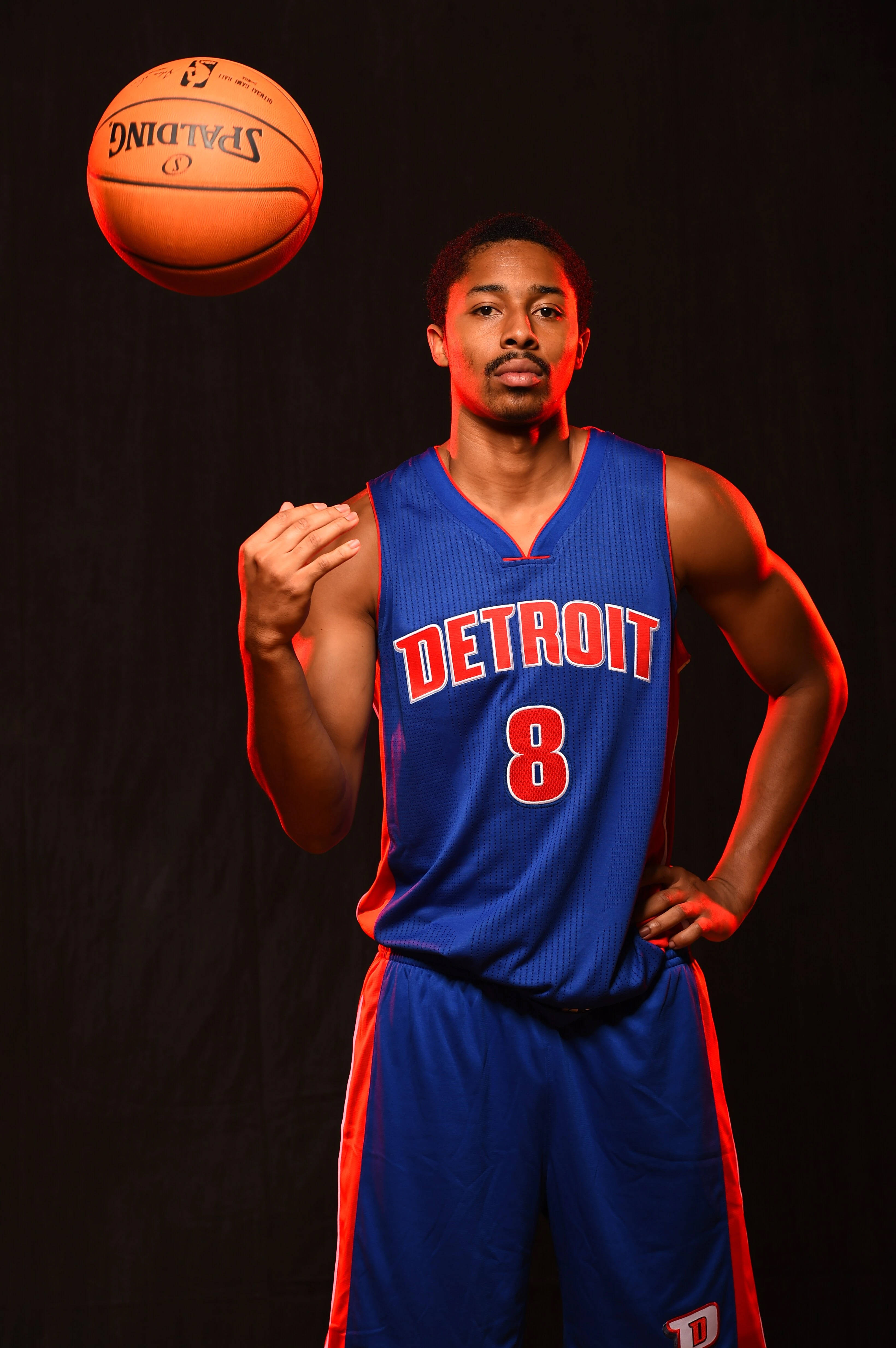 spencer dinwiddie 桌布 壁紙 — 一名身穿底特律活塞隊球衣的籃球運動員在黑色背景和戲劇性紅色燈光下平衡一個Spalding籃球。