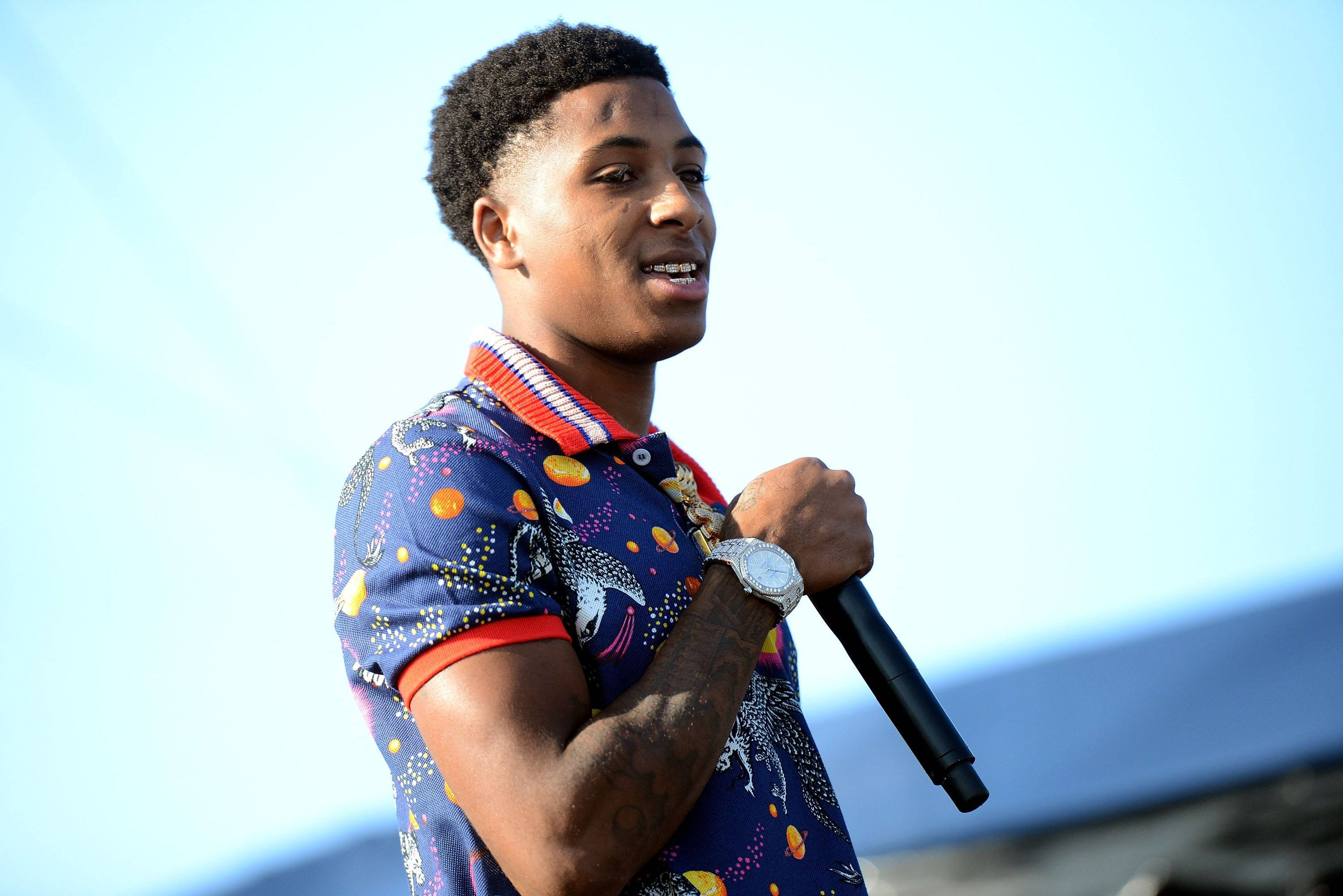 Youngboy 桌布 壁紙 — 一名身穿彩色圖案襯衫的年輕男子在明亮的藍天下戶外表演時手持麥克風。