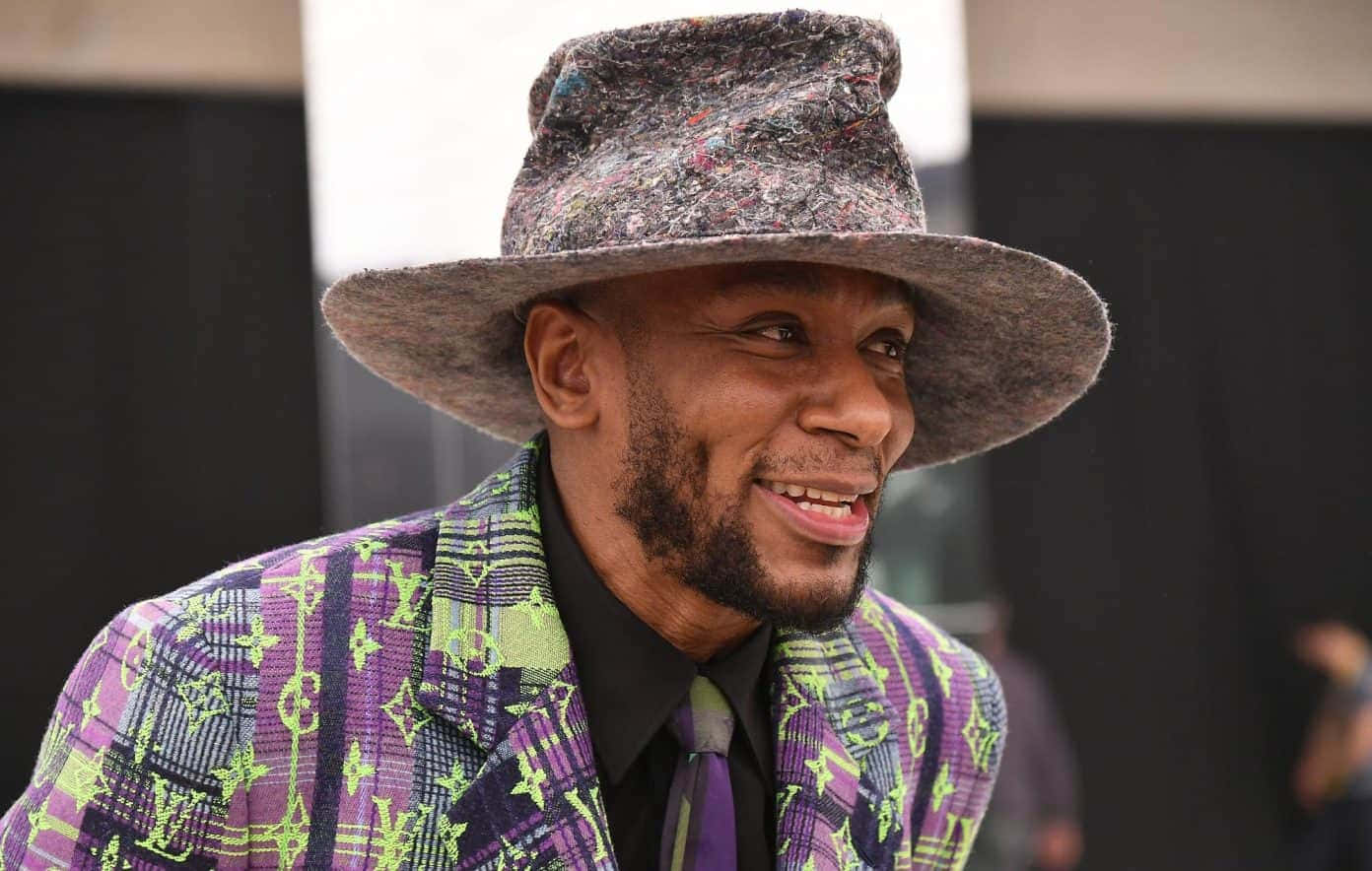 Yasiin Bey 桌布 壁紙 — 一名身穿彩色圖案西裝和斑點帽子的男子在活動中溫暖地微笑。