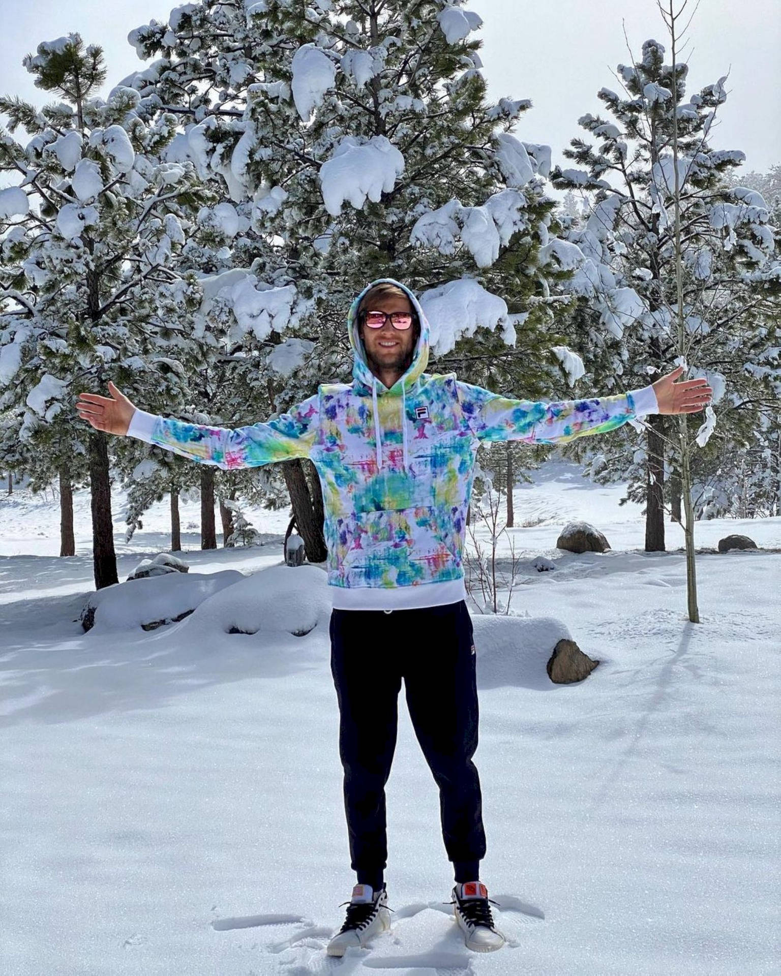  壁紙 — 一名身穿彩色紮染連帽衫的男子在白雪覆蓋的松樹林中，開心地張開雙臂站立。