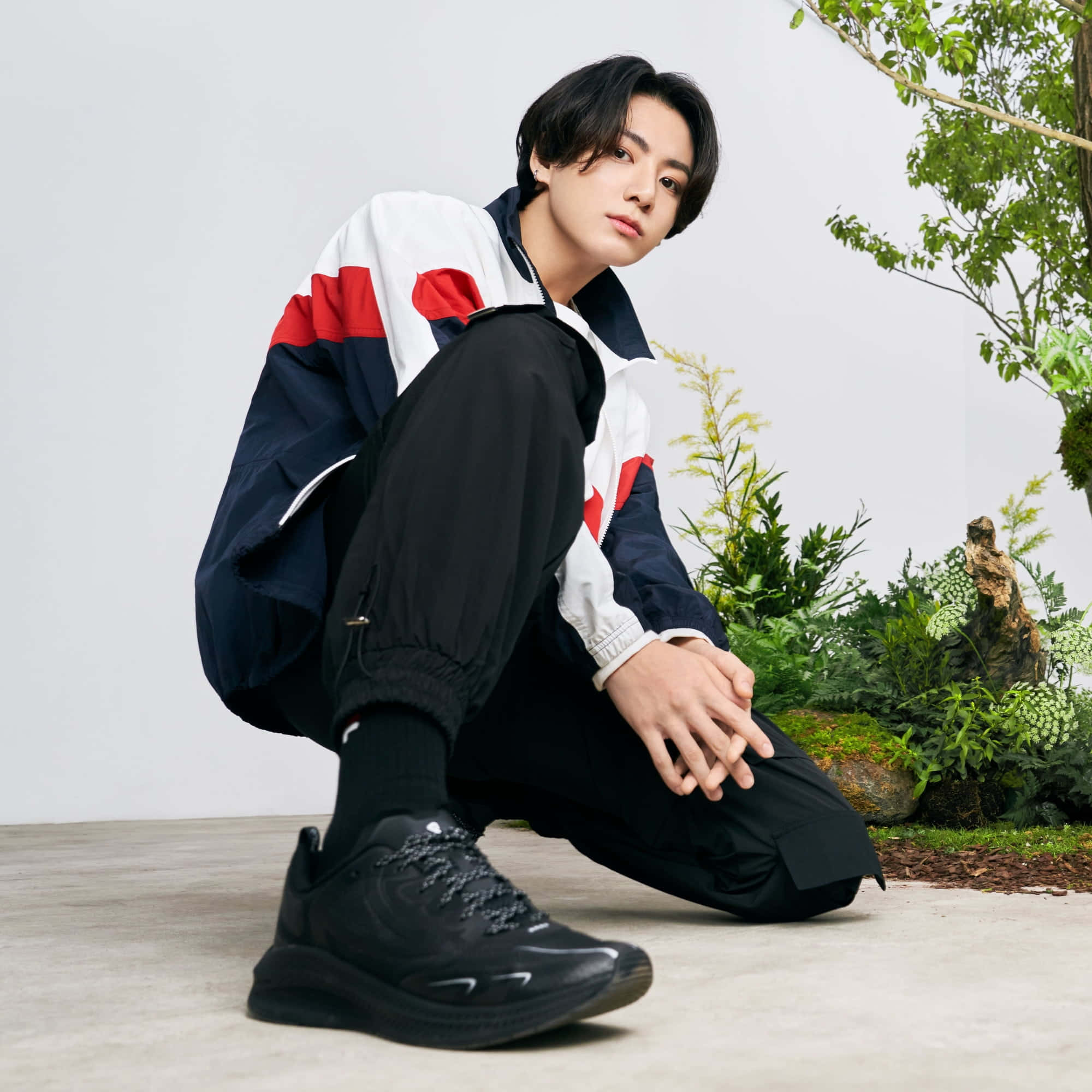 Jungkook 桌布 壁紙 — 一名身穿彩色運動外套的年輕男子蹲在茂密綠植旁，背景是純白牆壁。