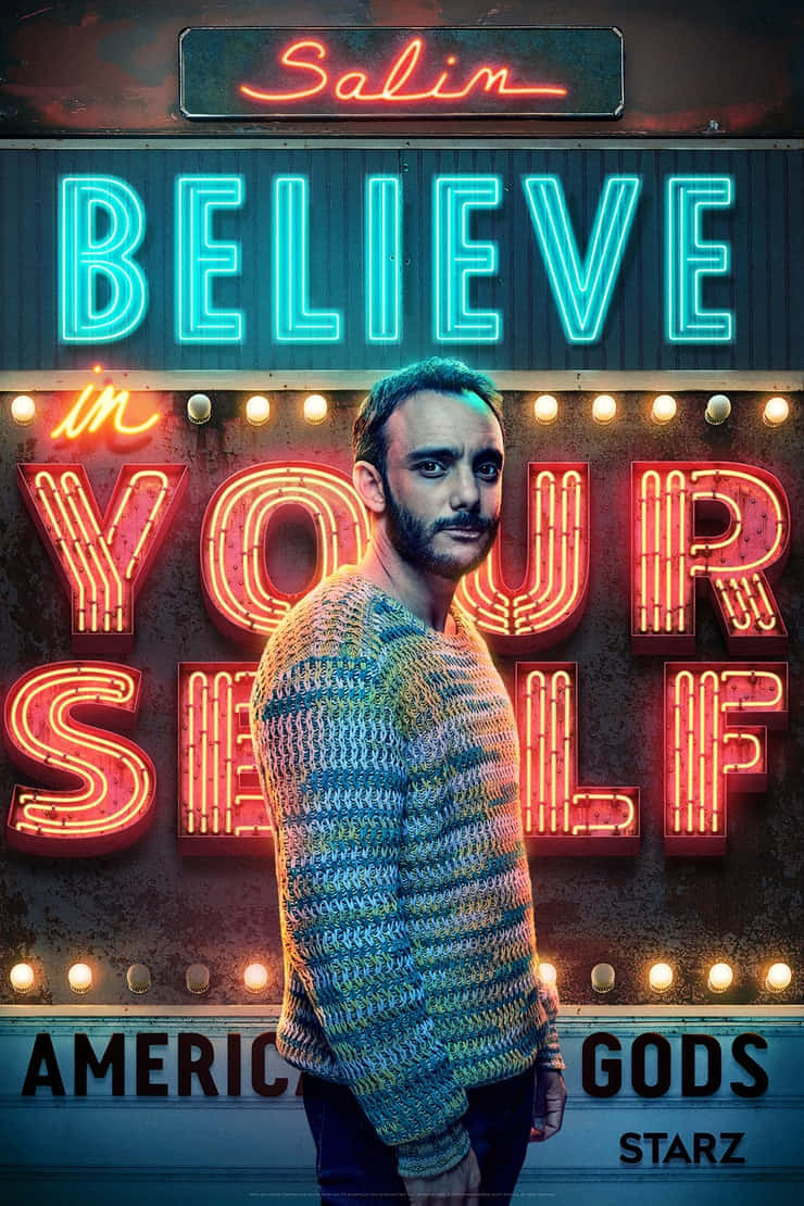 American Gods 桌布 壁紙 — 一名身穿彩色針織毛衣的男子站在一個寫有「Believe in Yourself」並帶有「American Gods」品牌的鮮豔霓虹燈牌前。