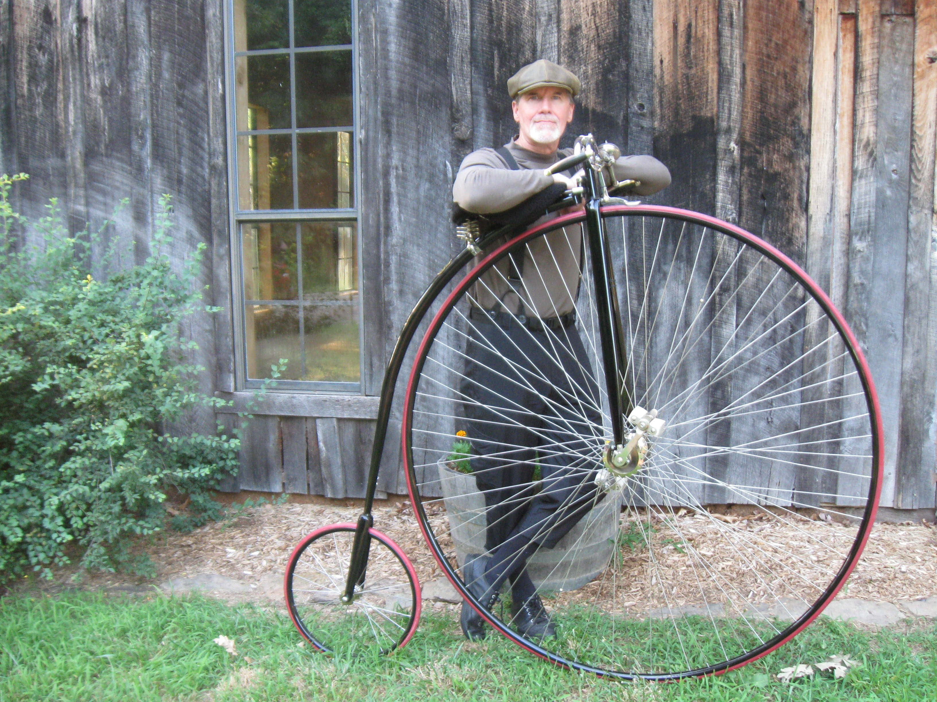 Bicycle 桌布 壁紙 — 一名身穿復古服裝的男子，自豪地站在一輛經典高輪腳踏車旁，背景是風化的木造建築。