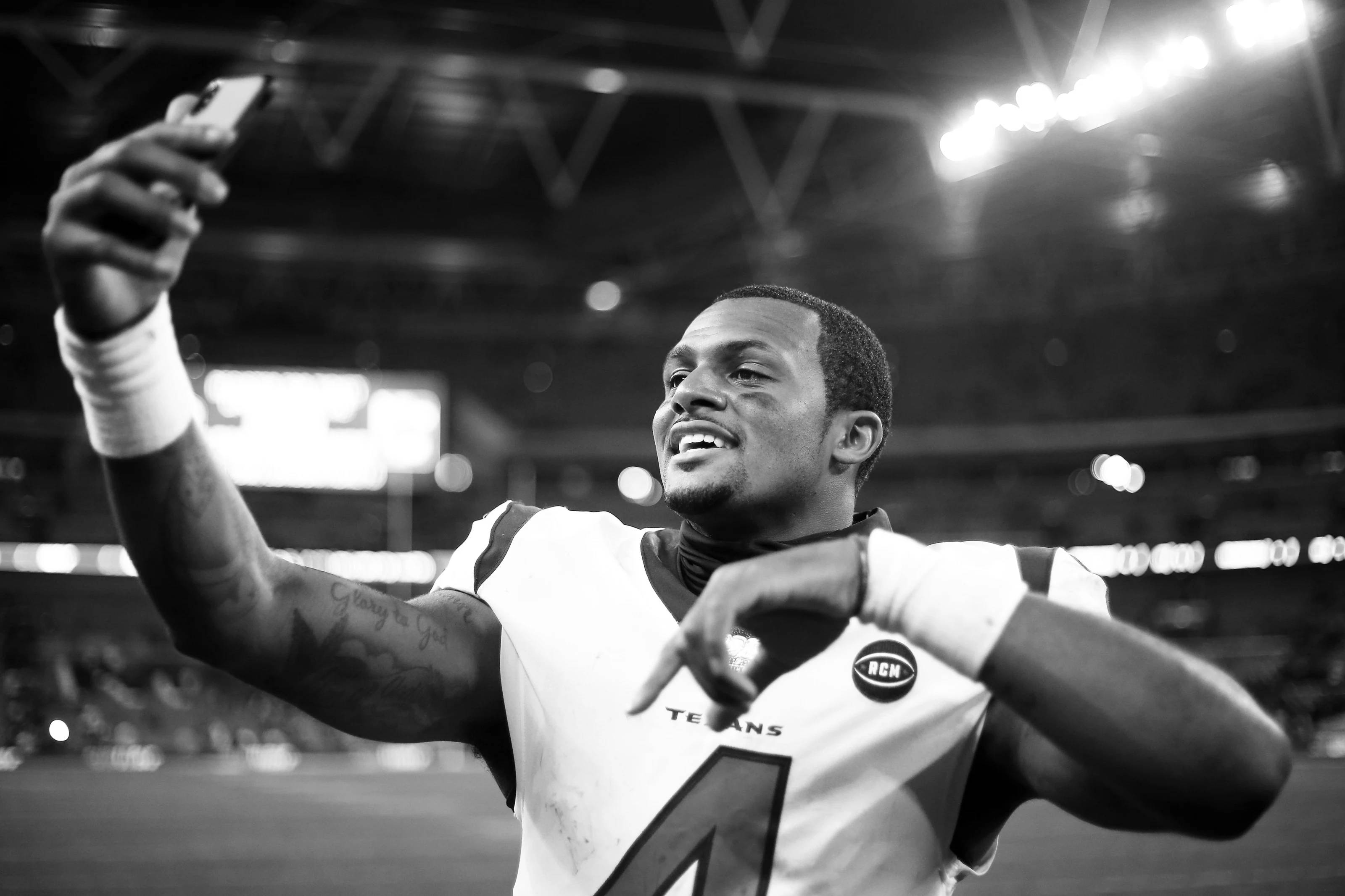 Deshaun Watson 桌布 壁紙 — 一名身穿德州人隊球衣的足球運動員在體育場燈光下，用手機自拍，露出燦爛的笑容。