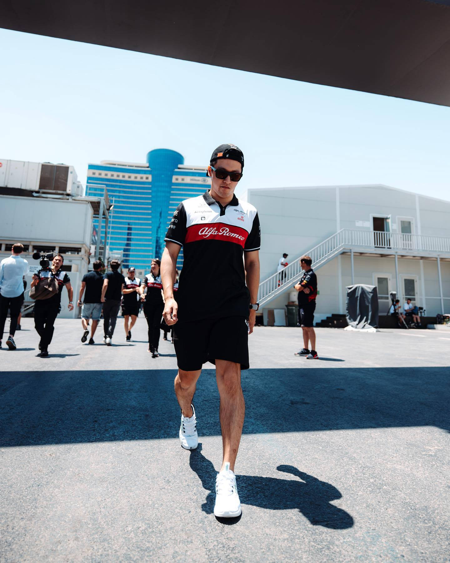 Sunglasses 桌布 壁紙 — 一名身穿愛快羅密歐賽車隊服裝的男子，在賽車場地明亮的陽光下自信地行走。