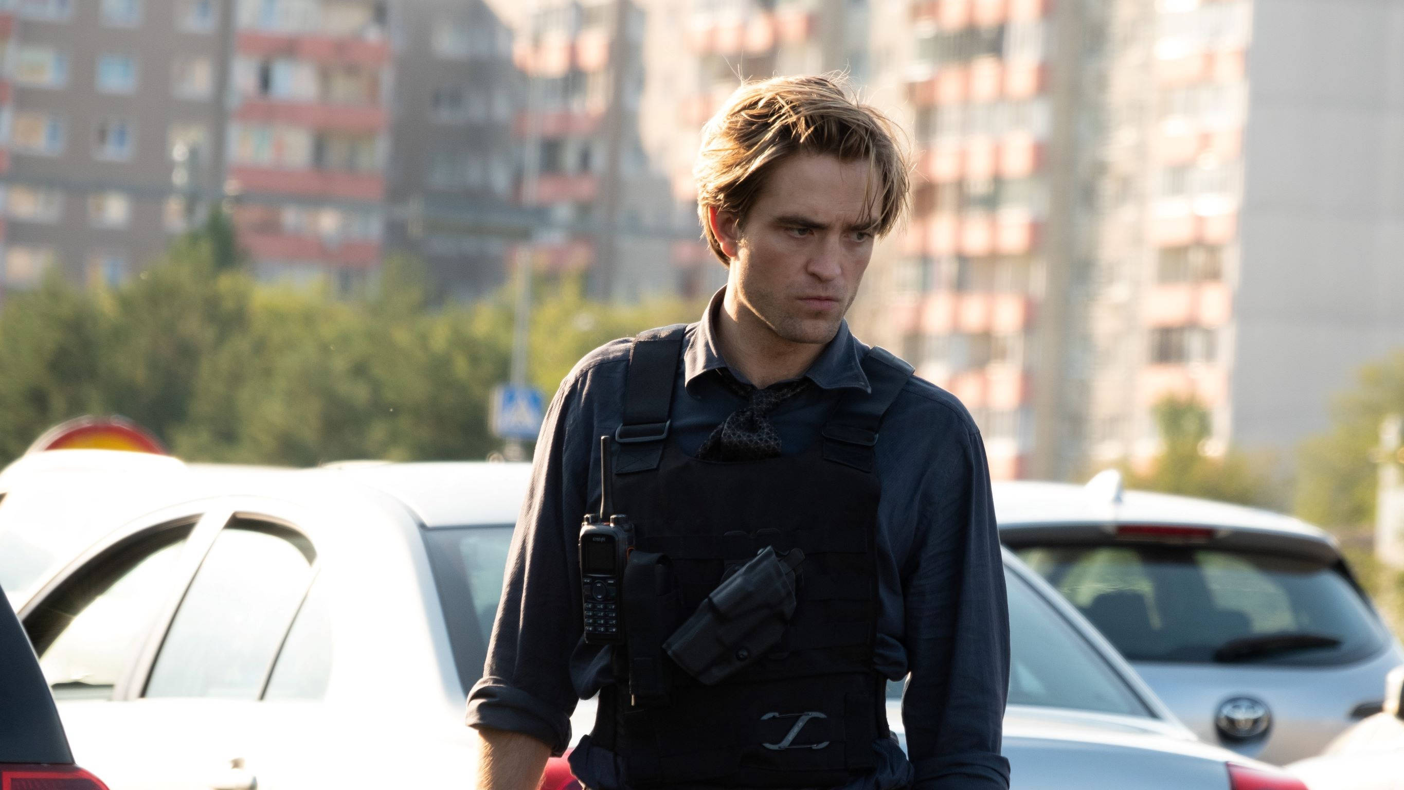 Robert Pattinson 桌布 壁紙 — 一名身穿戰術裝備的男子莊嚴地站在停泊的汽車之間，背景是公寓大樓。