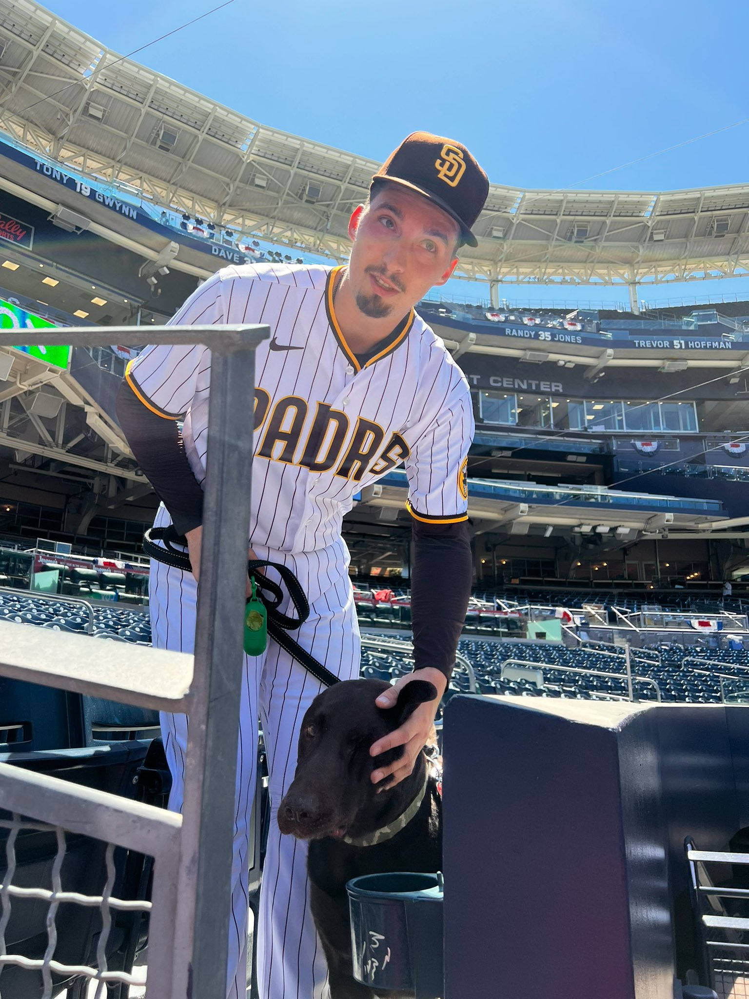 Blake Snell 桌布 壁紙 — 一名身穿教士隊球衣的棒球運動員在陽光明媚、座位空無一人、藍天白雲的球場上撫摸一隻黑狗。