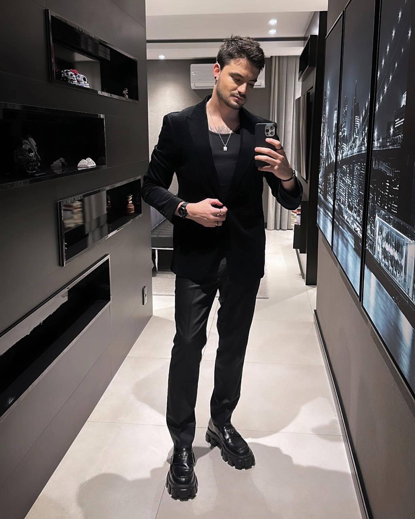 Mens Fashion 桌布 壁紙 — 一名身穿時尚黑色西裝和厚底靴子的男子，在一個現代簡約的走廊裡，對著鏡子自拍，牆上掛著城市景觀藝術品。
