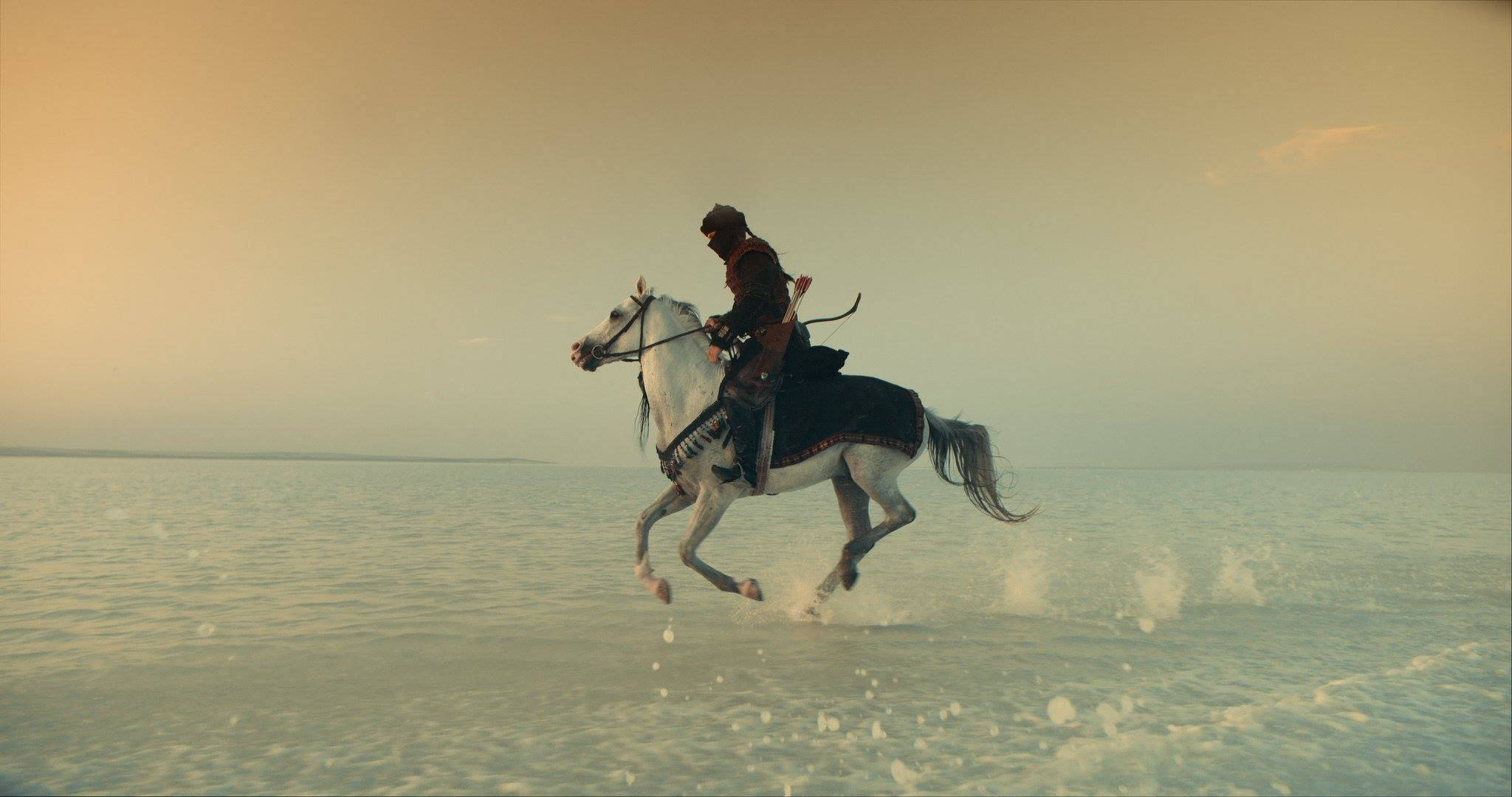 Ertugrul 桌布 壁紙 — 一名身穿暗色盔甲的騎士騎著白馬，在朦朧的金色天空下，奔馳於淺水之中。