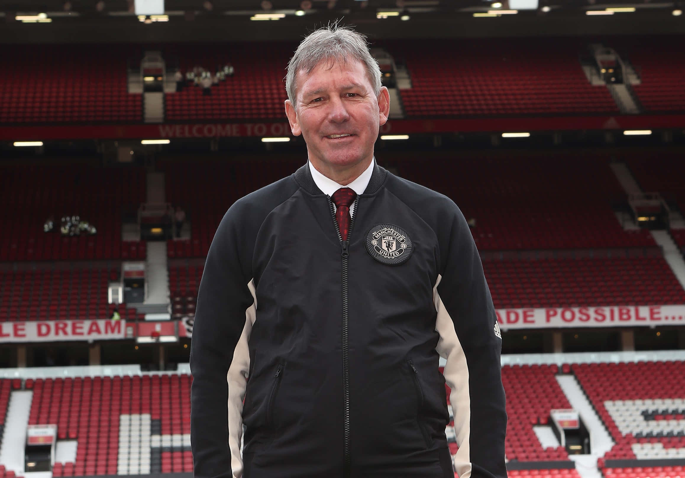 Smile 桌布 壁紙 — 一名身穿曼聯外套的男子在一個有紅色座位和'WELCOME TO OLD TRAFFORD'標誌的體育館內自信地微笑。