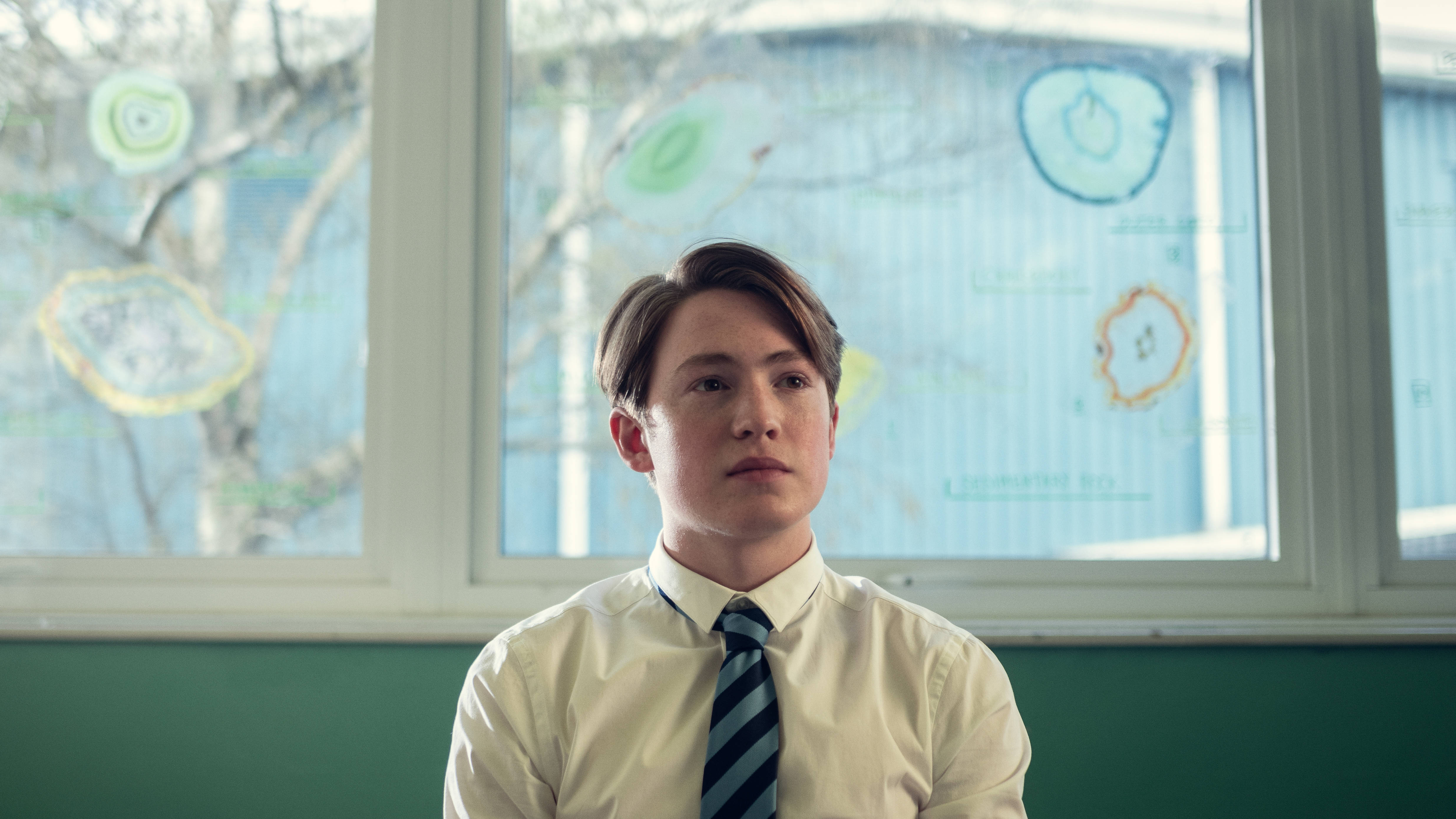 心碎男孩 桌布 壁紙 — 一名身穿有領襯衫和條紋領帶的年輕男子坐在教室裡，身後窗戶上有色彩繽紛的圖表，若有所思。