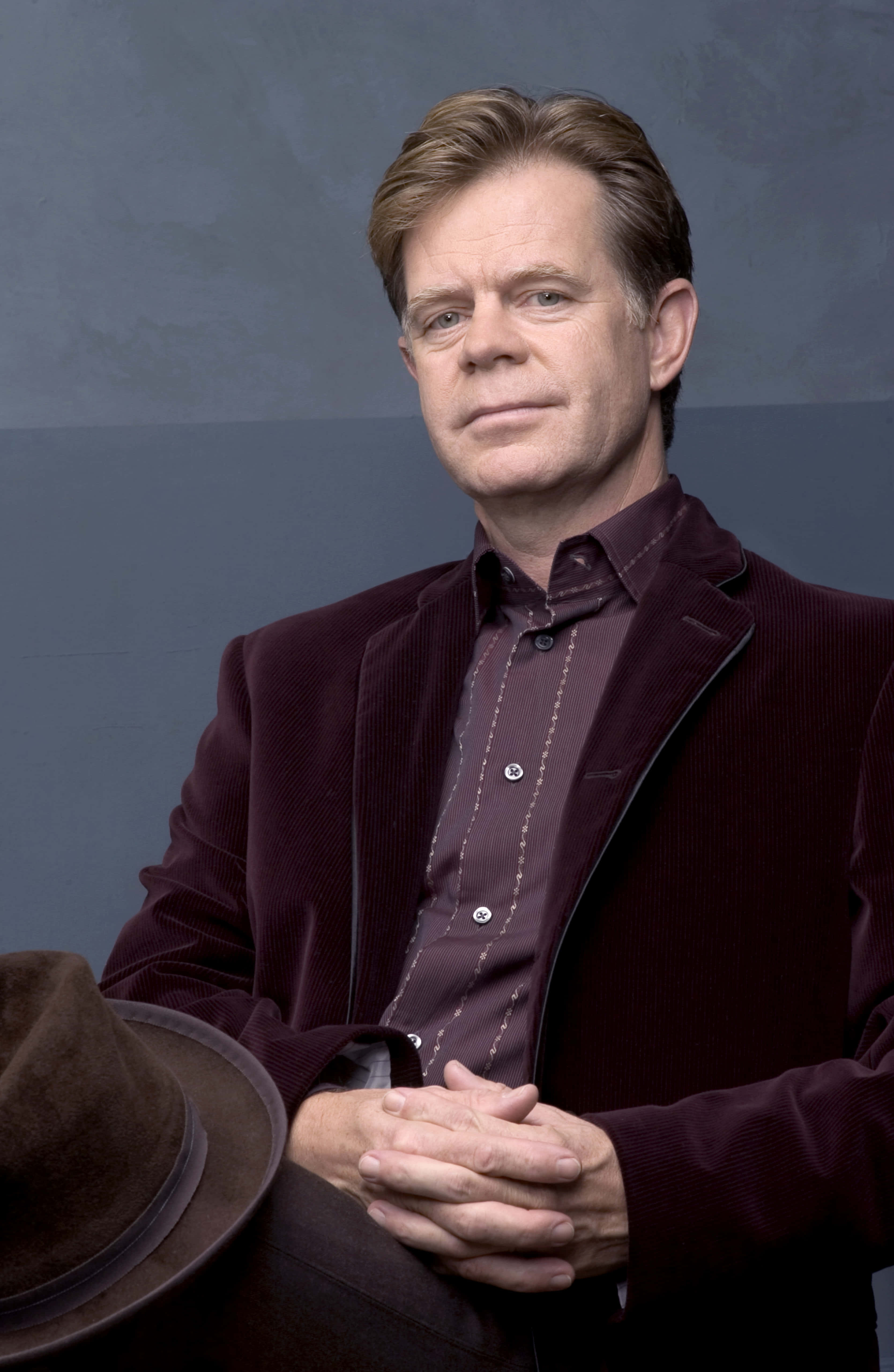william h. macy 桌布 壁紙 — 一名身穿栗色燈芯絨西裝及相配襯衫的男子，雙手交疊，靜靜地坐在一個棕色軟呢帽旁邊，背景為柔和的灰色。