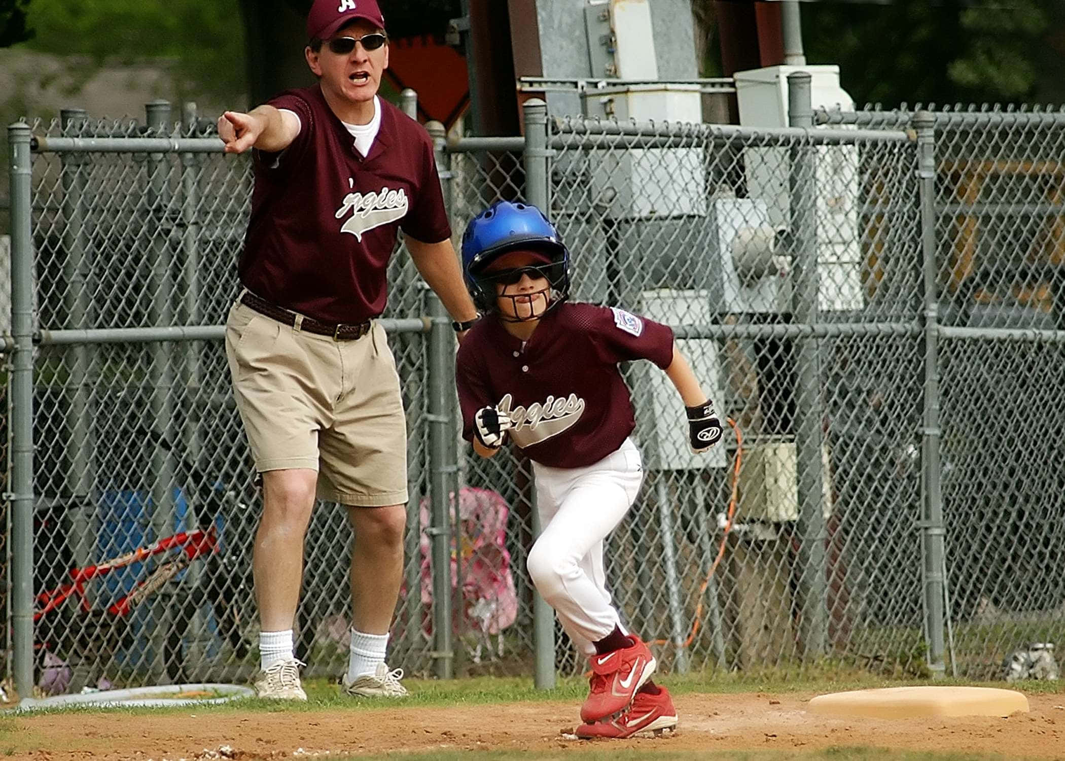 Little League Baseball 桌布 壁紙 — 一名身穿栗色球衣的年輕棒球選手正衝向壘包，教練在鐵絲網後方急切地做手勢。