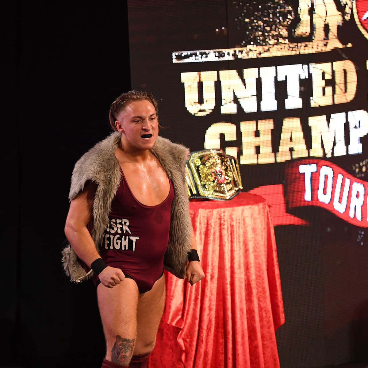 WWE 桌布 壁紙 — 一名身穿栗色緊身衣和毛皮背心的摔角選手，在明亮的舞台燈光下，勝利地站在紅色桌布覆蓋的桌子旁，桌上放著冠軍腰帶。