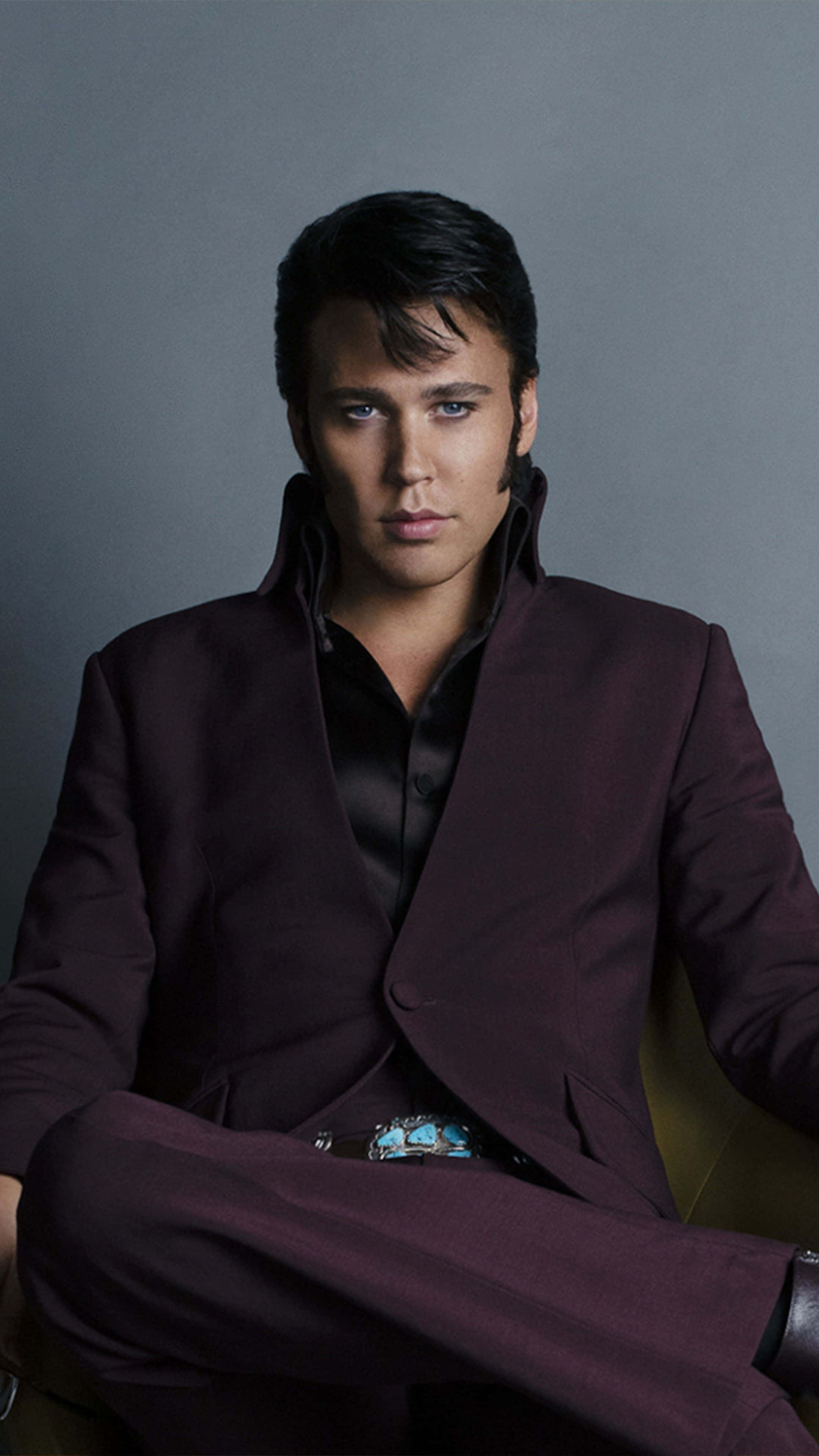 4K Elvis 桌布 壁紙 — 一名身穿栗色西裝、配戴綠松色皮帶扣的男子，自信地坐在灰色背景前。