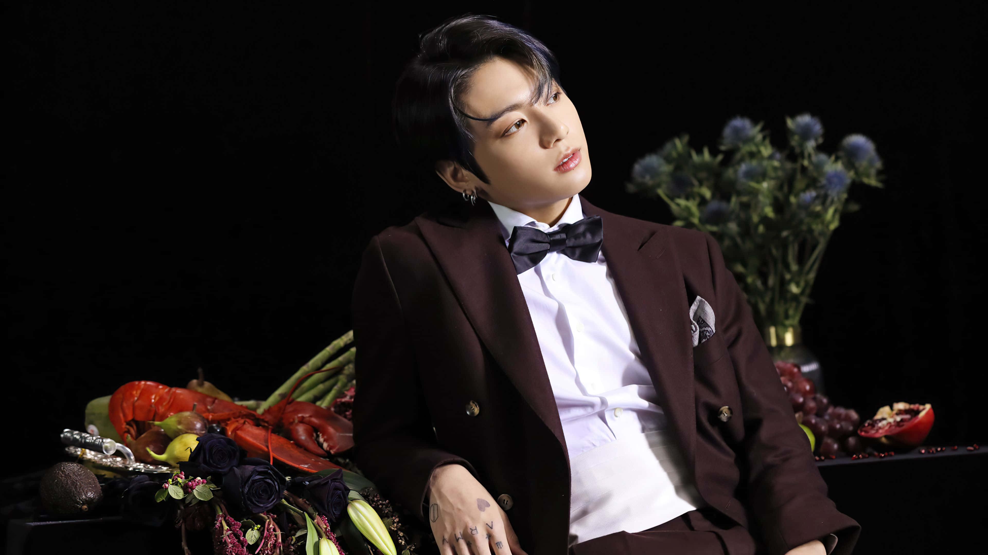 Jungkook 桌布 壁紙 — 一名身穿栗色西裝和領結的男子在黑色背景前優雅地擺姿勢，周圍環繞著鮮豔的花朵和水果。