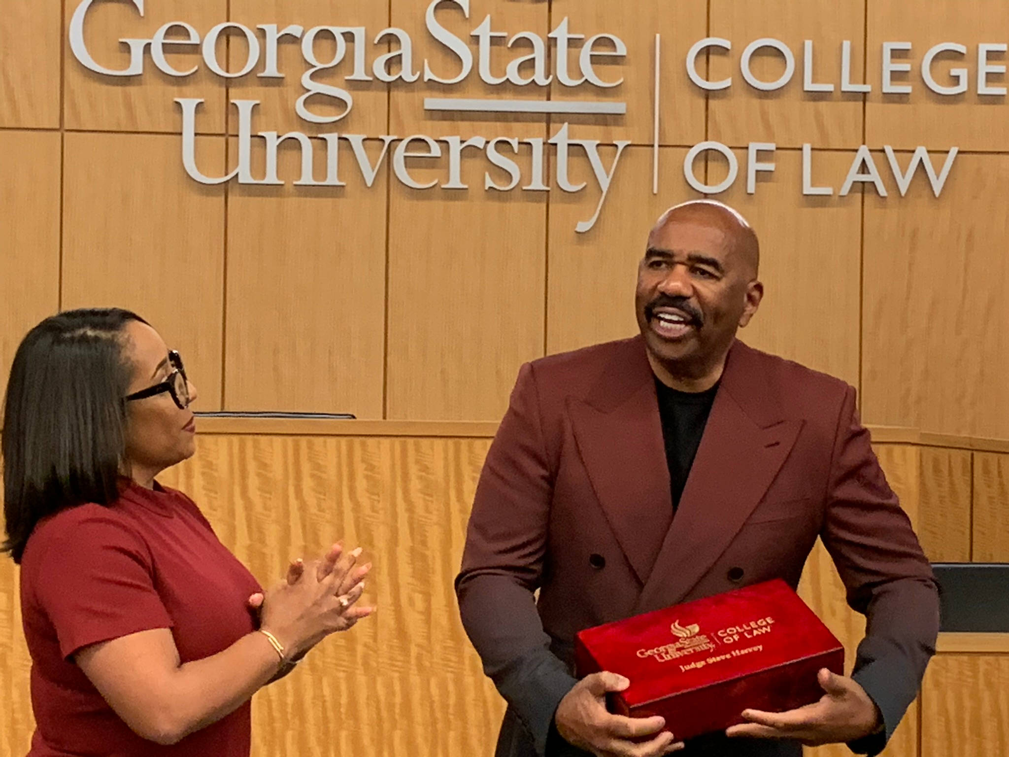 steve harvey 桌布 壁紙 — 一名身穿栗色西裝的男子手持紅色法學院榮譽盒，旁邊一名身穿紅色衣服的女子在喬治亞州立大學法學院鼓掌。