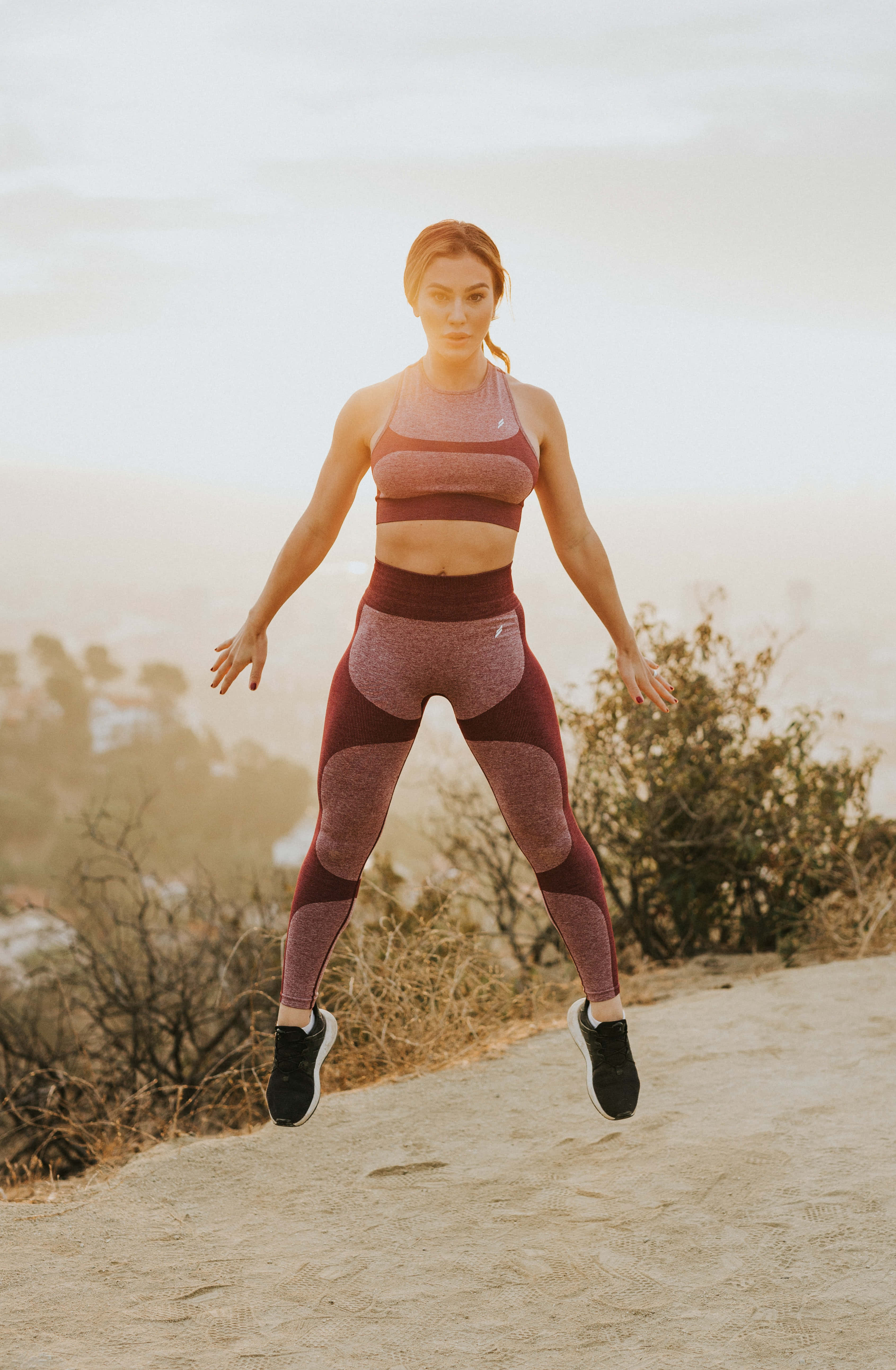 Exercise 桌布 壁紙 — 一名身穿栗色運動服的女性在塵土飛揚的小徑上躍起，身後是金色時刻中朦朧的城市天際線。