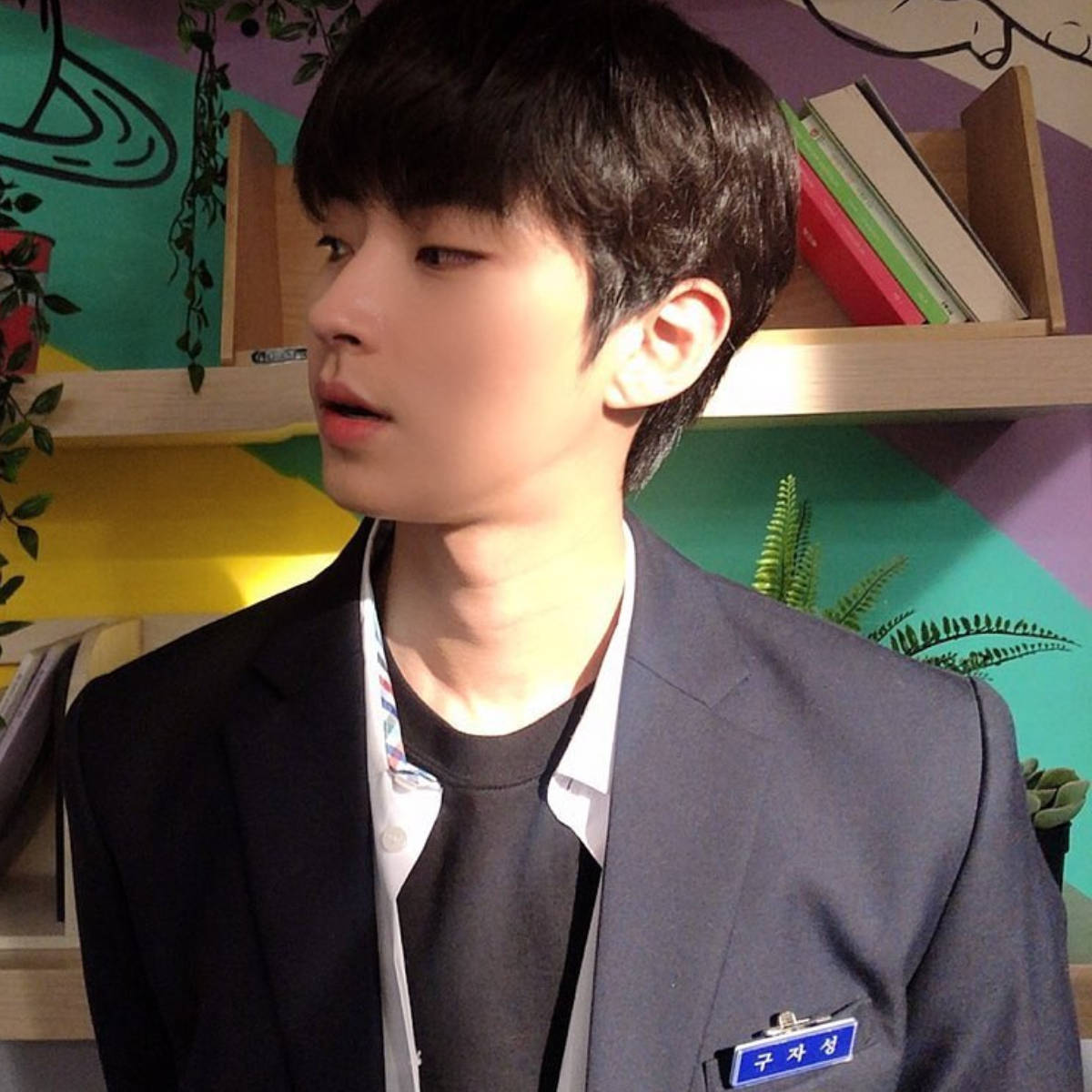 Hwang In Yeop 桌布 壁紙 — 一名身穿校服、帶著名牌的年輕男子站在一個色彩繽紛、有書架和植物的房間裡。