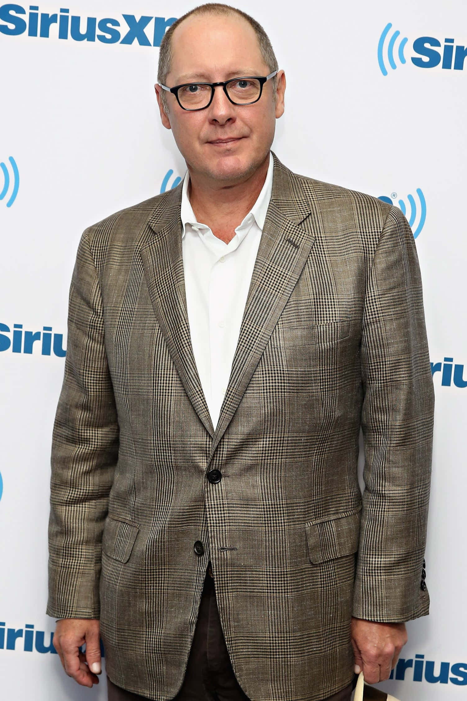 james spader 桌布 壁紙 — 一名身穿格紋西裝外套、戴眼鏡的男子自信地站在SiriusXM品牌背景前。