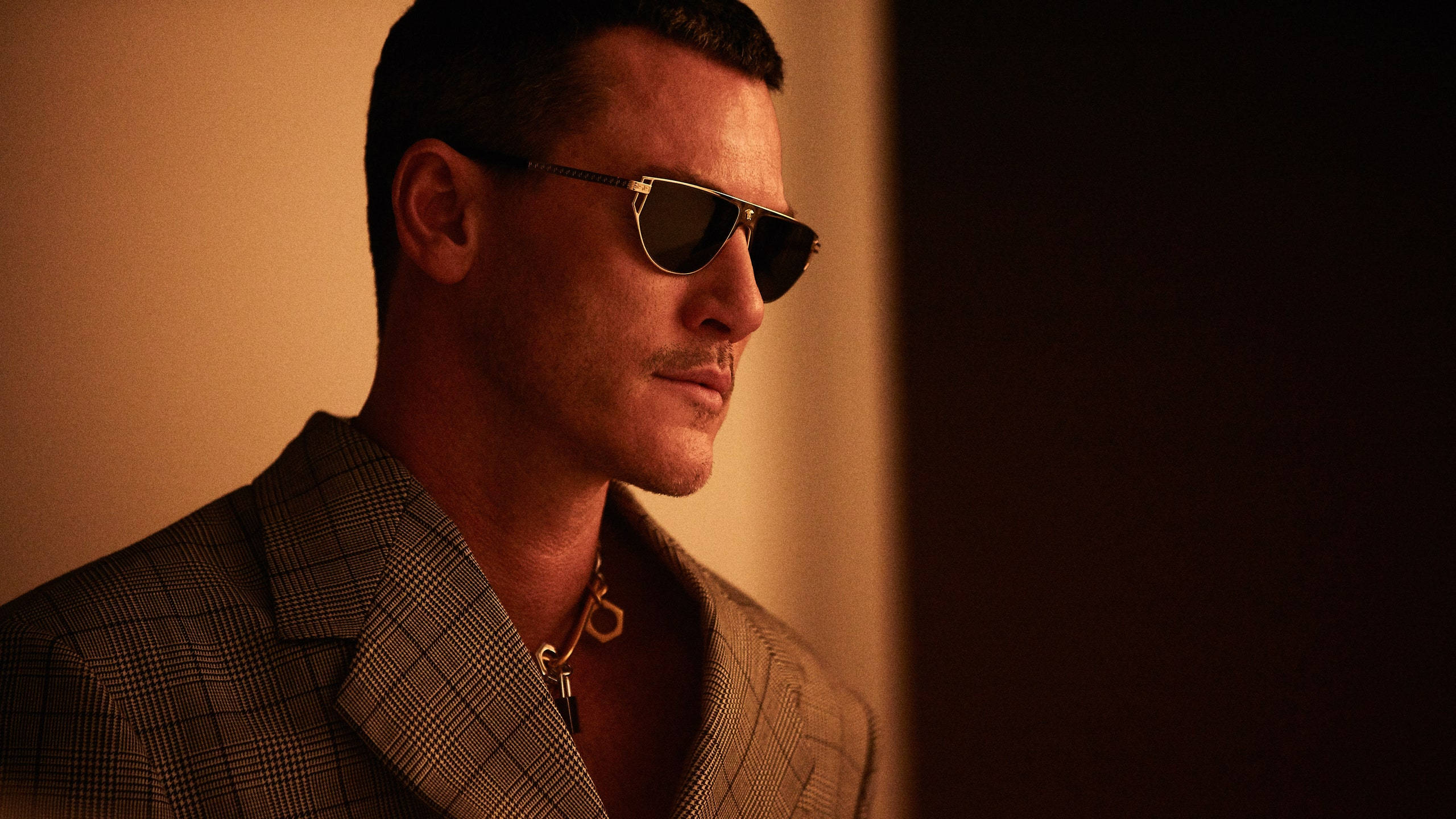 Sunglasses 桌布 壁紙 — 一名身穿格紋西裝外套、戴著飛行員太陽眼鏡的男子，在溫暖的金色光線下站在深色背景前。