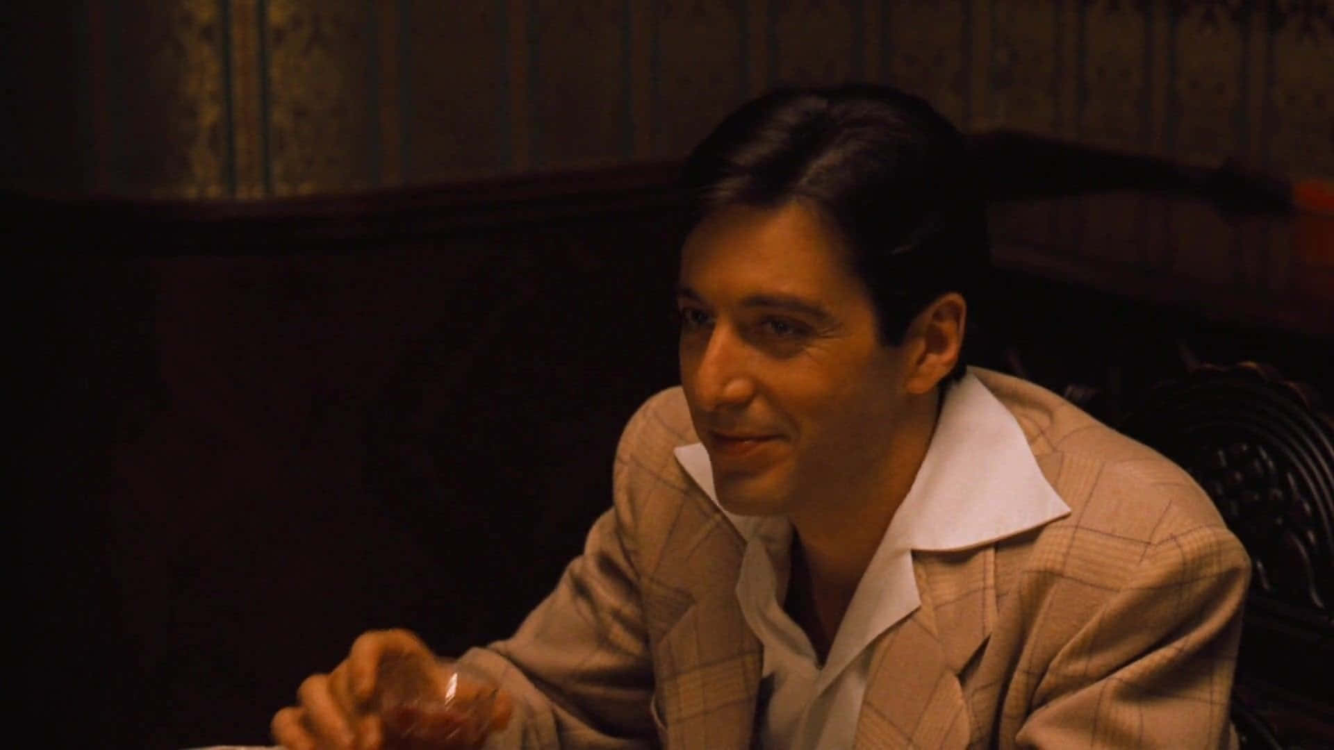 The Godfather 桌布 壁紙 — 一名身穿格紋西裝的男子在光線昏暗的復古房間裡，手持玻璃杯，對著花紋壁紙溫柔微笑。